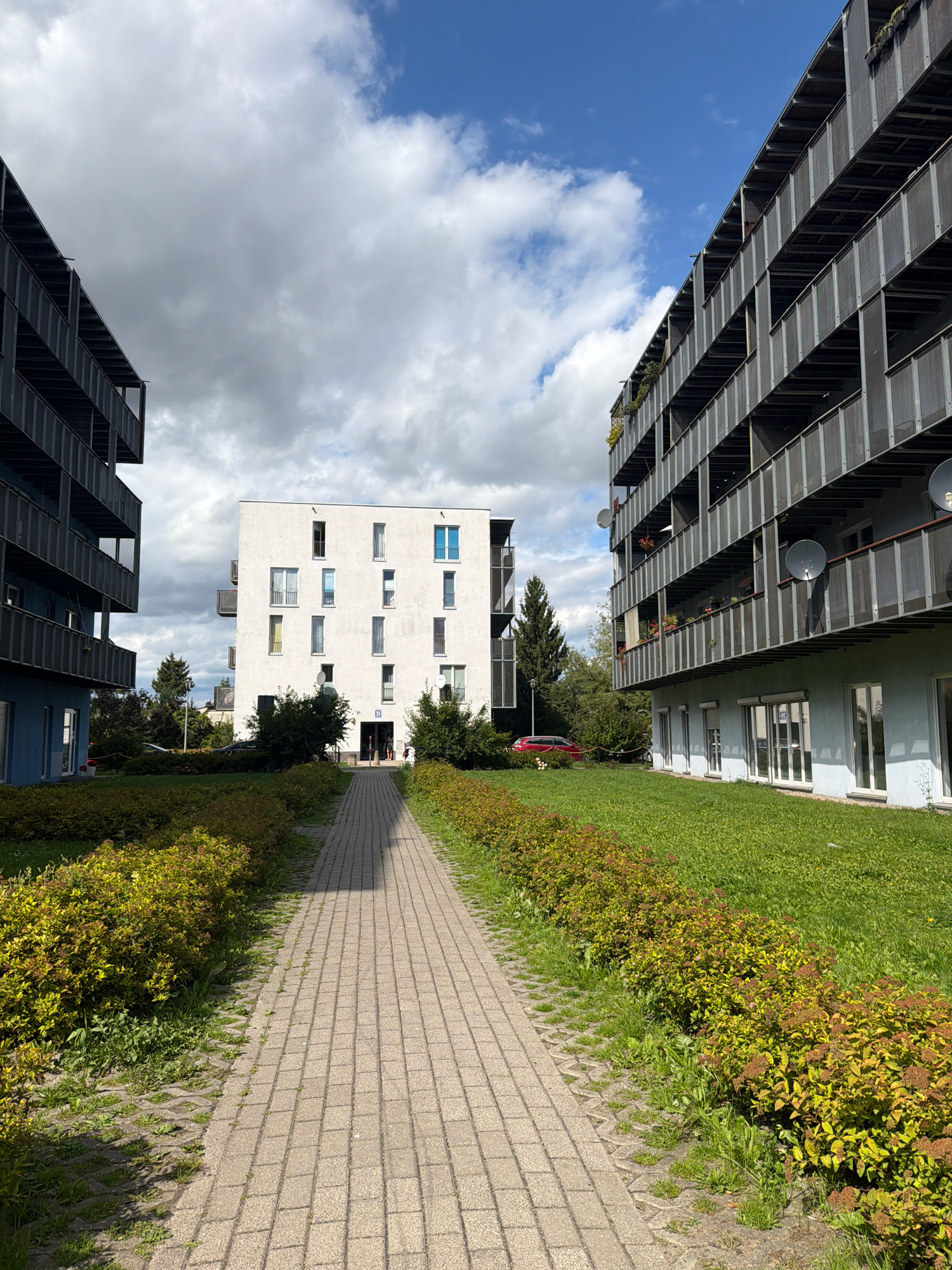 An outdoor shot features a paved walkway leading through a grassy area with manicured bushes towards a modern, white, multi-story apartment building. The building has numerous windows and balconies, and a sign with the number "35" is visible near the entrance. To the left and right of the walkway, two other apartment buildings are visible, one appearing darker and more industrial with metal railings on the balconies, and the other a lighter blue on the lower levels. The sky is partly cloudy with a mix of white clouds and blue sky. A red car is parked on the grass to the left of the walkway, near some trees. In the foreground, the walkway is bordered by dense, green bushes with some reddish-brown elements. The scene appears to be in a residential area. This image was taken in Koszalin, Poland.