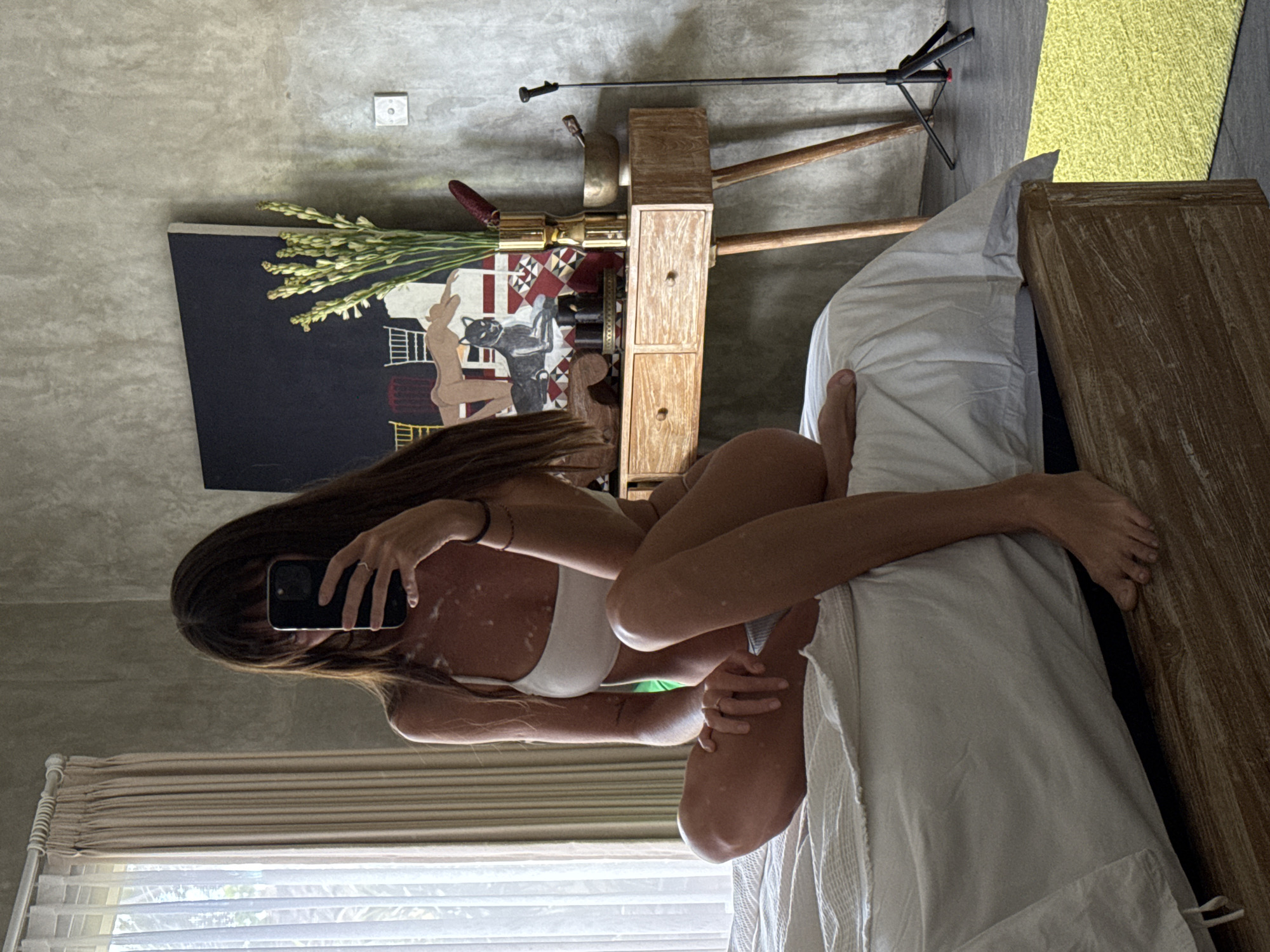 A woman with long brown hair is taking a selfie in a mirror. She is wearing a white bikini top and green bottoms. She is holding a black smartphone in her left hand and looking into the screen. She is sitting on a white bed. Behind her are partially drawn white curtains and a window. On the wall are a dark painting, a wooden cabinet, and a black tripod. This image was most likely taken in an accommodation in North Kuta, Indonesia.