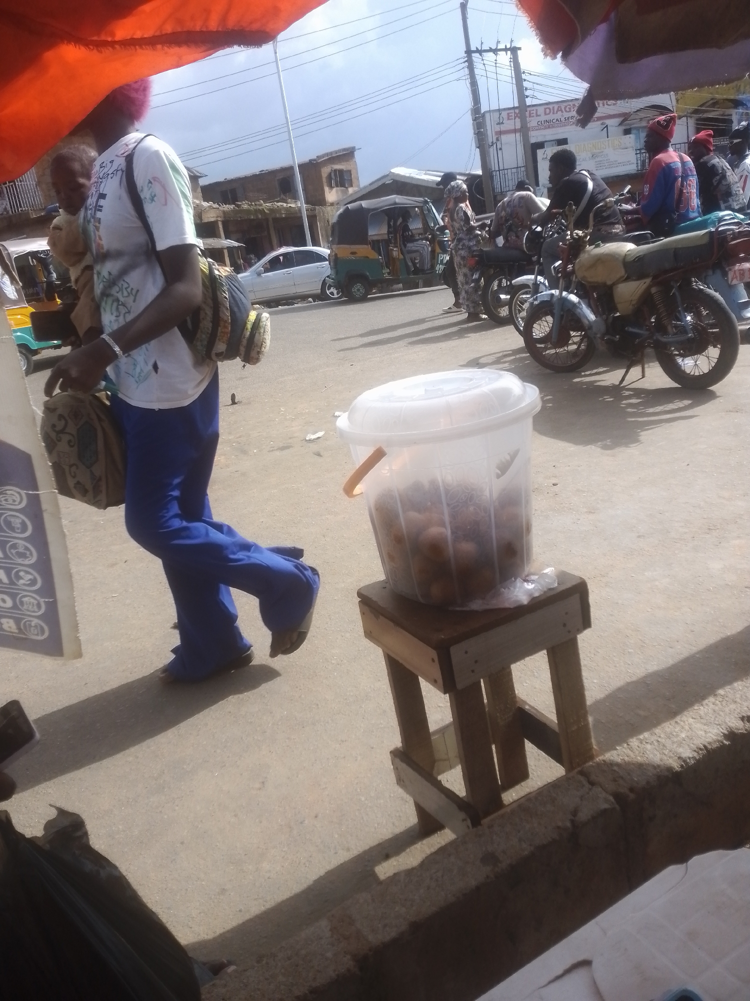 In an outdoor, likely urban setting, a person with pink-dyed hair walks by carrying a bag over their shoulder and another in their hand. They are wearing a white t-shirt with graffiti-style lettering and blue pants. A transparent plastic bucket filled with small, round objects sits on a wooden stool in the foreground. The scene appears to be a busy street, with several motorcycles parked and people gathered around them. In the background, buildings and a sign that reads "EXCEL DIAGNOSTICS CLINICAL SERVICES" are visible. The lighting suggests it is daytime, possibly mid-morning or afternoon, under a partly cloudy sky. The overall impression is of a bustling marketplace or a busy street with roadside vendors.