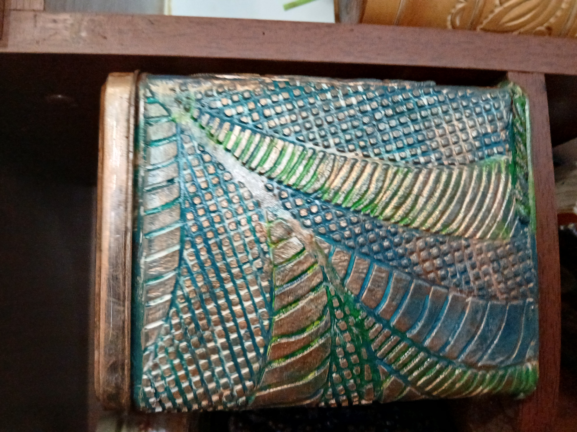 A close-up, eye-level shot shows a textured, decorative tin with a metallic gold lid. The tin is predominantly teal and gold, with a raised pattern that mimics woven or scaled textures. The pattern is highly detailed and appears to be a stylized representation of leaves or feathers. The tin is positioned on a dark wooden shelf, with another similarly textured object partially visible to the right. The background is dark and out of focus, suggesting an indoor setting. The lighting is ambient and diffused. The image does not contain any recognizable landmarks or indicators of a specific city like Lipetsk, Russia.