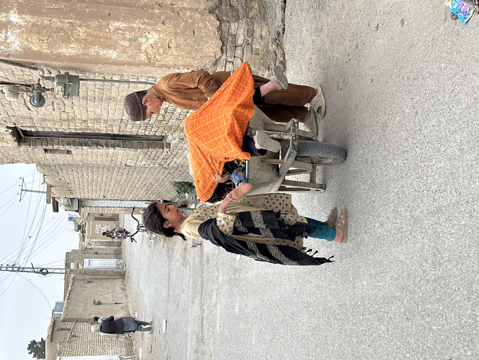 The image depicts a street scene in Quetta City Tehsil, Pakistan, under an overcast sky. In the foreground, three individuals, appearing to be children, are positioned around a grey metal wheelbarrow. A young girl, dressed in a cream-colored tunic with colorful polka dots, teal trousers, and tan sandals, with a black patterned shawl draped over her left shoulder, stands on the left. She is holding the bare foot of a child who is largely obscured inside the wheelbarrow. This child is covered by a bright orange fabric with a grid pattern; a second foot, wearing a light grey slipper, is visible over the right edge of the wheelbarrow. To the right of the wheelbarrow stands a young boy, wearing a brown kurta and a brown skullcap, with light grey slip-on sandals. The ground is a grey paved surface. The background features weathered buildings constructed of light-brown brick and mud-brick, lining both sides of the street. A utility meter and associated piping are affixed to the building on the right. Overhead utility poles and a dense network of electrical wires are visible against the pale sky. In the distance, an individual carrying a child walks away on the left side of the street, and another individual is visible on a motorcycle further down the thoroughfare. A single dark tree is present on the distant left horizon.