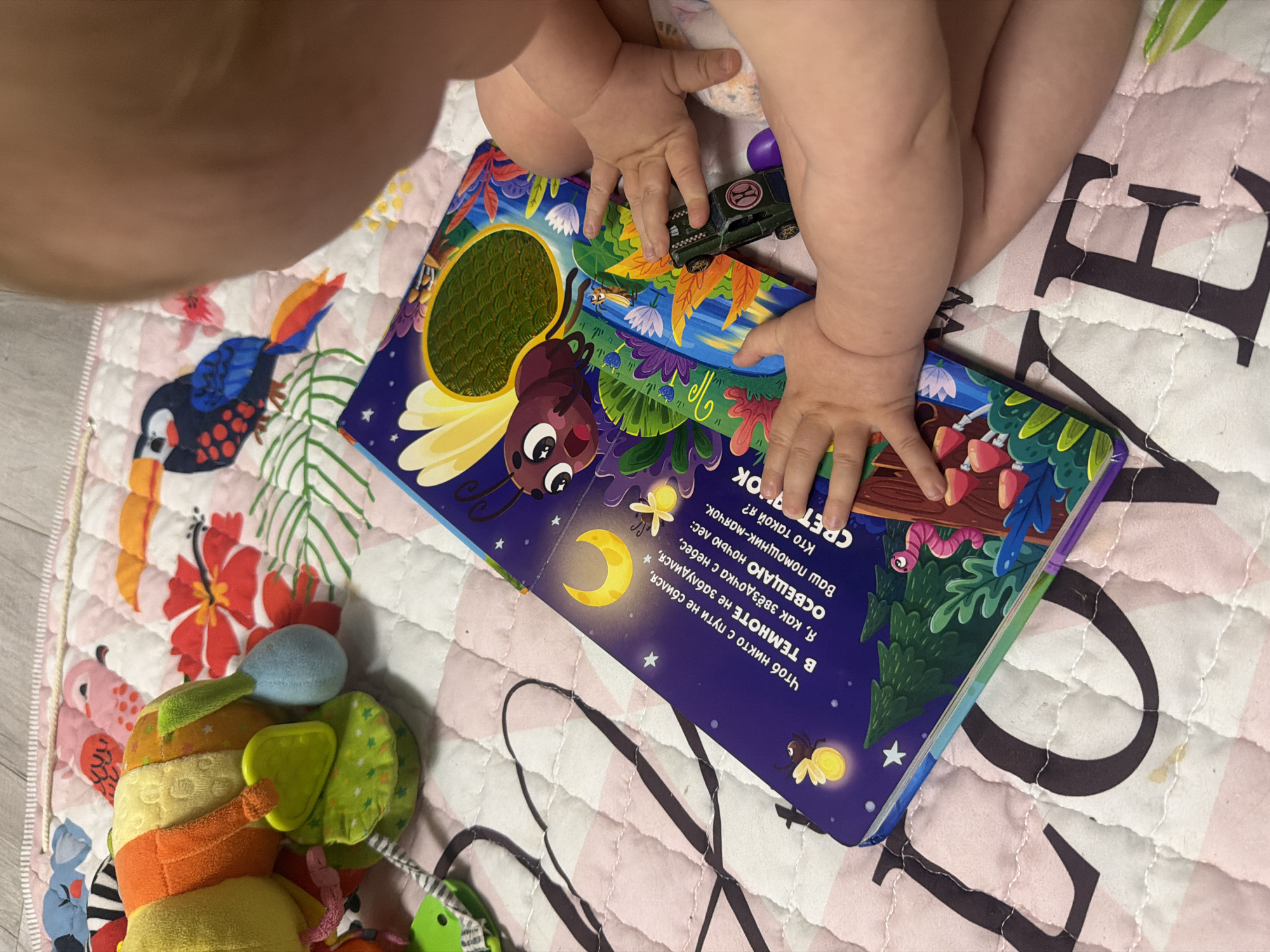 An overhead, medium shot captures a close-up of a baby's hands interacting with an open, colorful children's book lying on a patterned play mat. The book, featuring illustrations of a moth, a crescent moon, and fireflies, is positioned at an angle in the lower half of the frame. The baby's fingers are touching the page, and a small, dark toy car is placed on the book's spine. To the left of the book, a soft, plush caterpillar toy with various colored segments is visible. The play mat has a pink and white quilted design with large black letters, a portion of which appears to spell "LOVE." The background shows a light wooden floor. The image is taken indoors, likely in a home environment. The location of Rostov-on-Don, Russia is not evident from the visual information in the image.