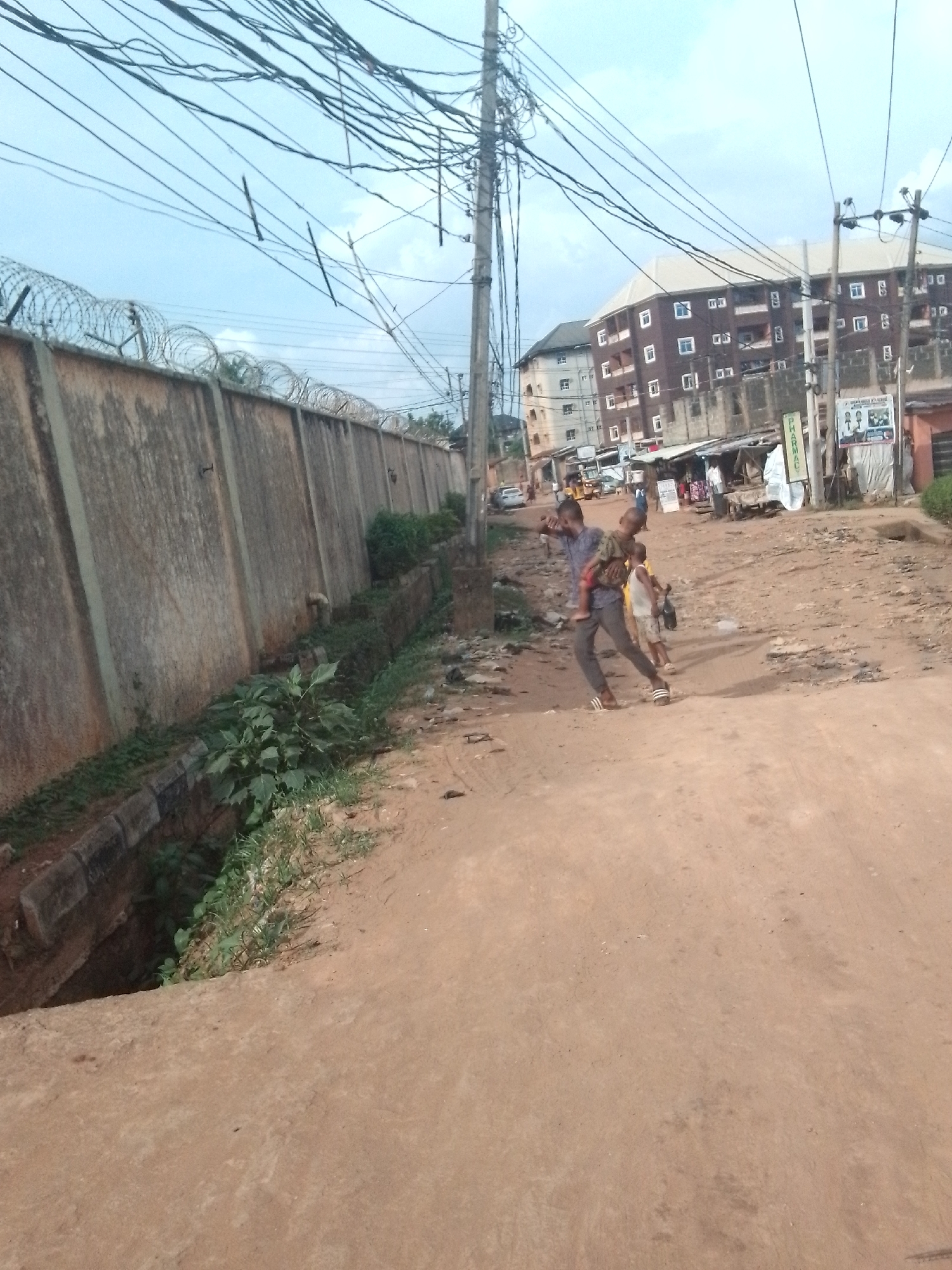 The image shows a street scene in Obosi, Nigeria. A man is carrying a child, with another child walking alongside. Buildings of several stories are visible in the background, along with a row of small shops or stalls. Overhead are numerous electrical wires. A high concrete wall topped with barbed wire runs along one side of the street, bordered by a drainage ditch. The road itself is unpaved and appears rough.