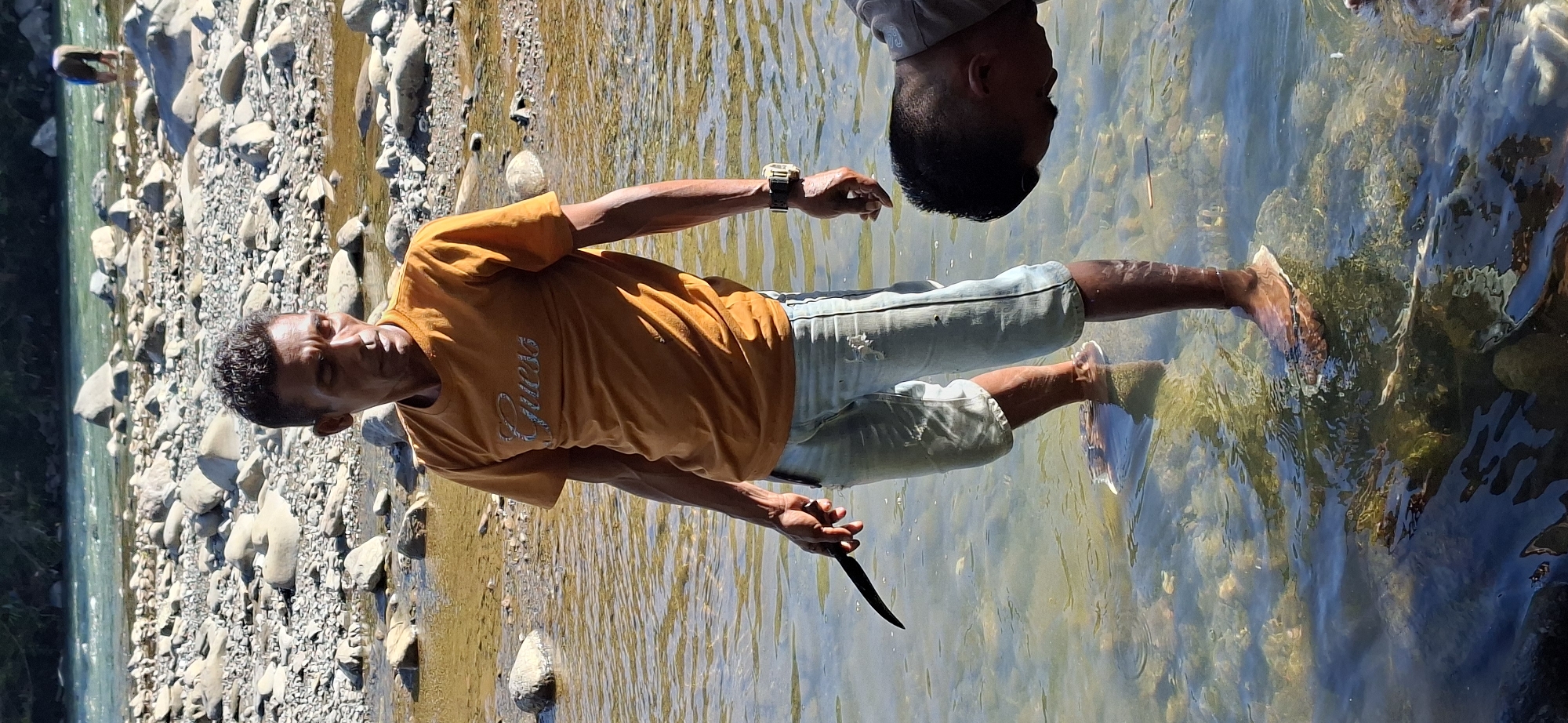 The image depicts an outdoor scene likely in or near Mane, Indonesia, characterized by a river or shallow body of water with a rocky bed. In the foreground, a man stands in the water, wearing an orange t-shirt with "Guess" printed on it, light blue distressed denim shorts, and sandals. He is holding a dark-colored knife in his left hand. His posture suggests he is engaged in some activity in the water. To the right of the frame, another person's head and shoulders are visible, bent down and looking into the water, suggesting a shared activity or interest. The background shows more of the rocky riverbed and a lush, tree-covered hillside under a bright sky, indicating a natural, possibly rural, environment. The lighting suggests it is daytime, with sunlight reflecting off the water's surface.