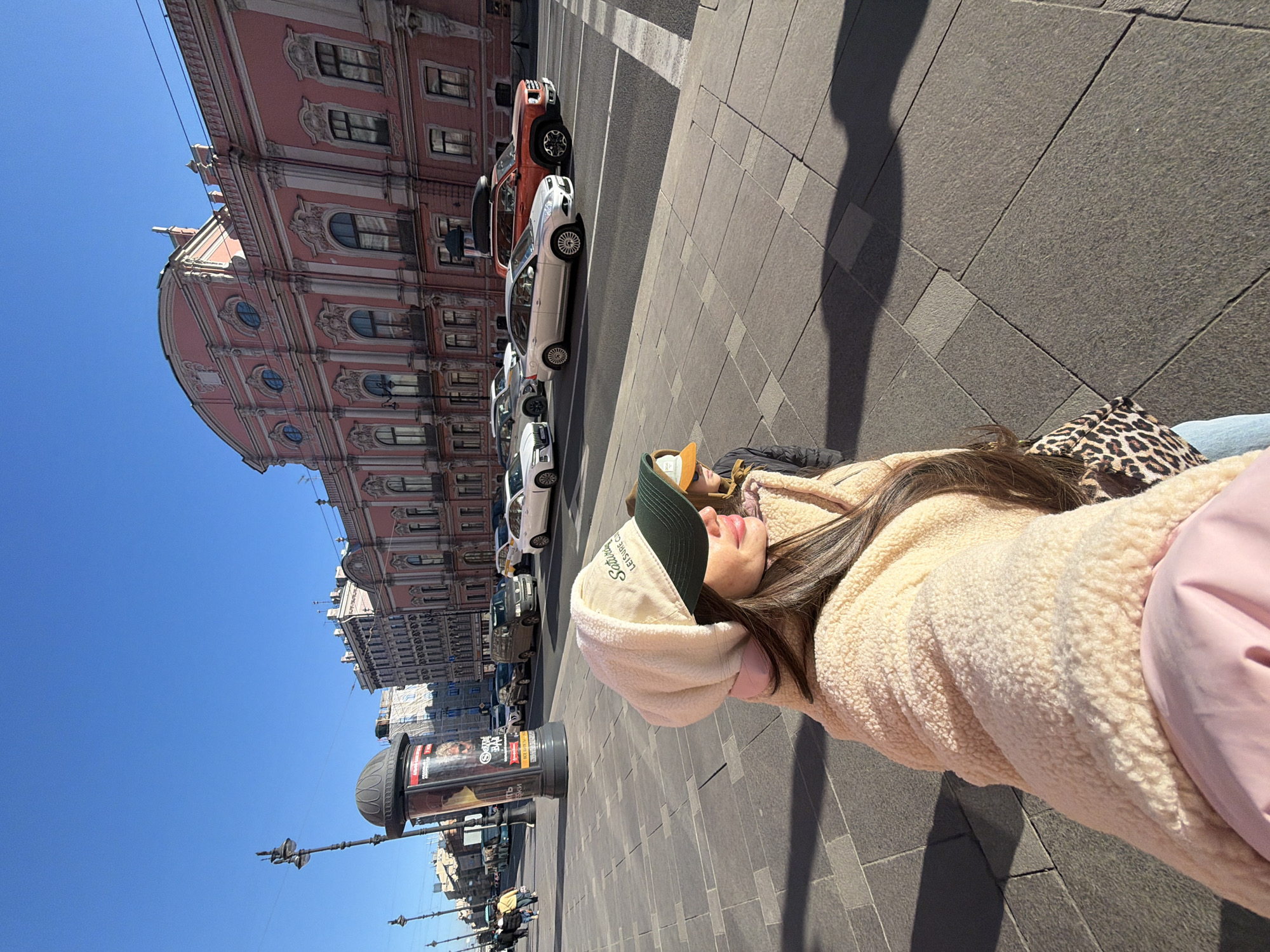 The image shows a person wearing a beige sherpa jacket, pink shirt, and a beige and green cap, lying on a grey brick sidewalk. The person has long brown hair and is partially visible. The background features the urban environment of Saint Petersburg, Russia. A line of cars is parked along the street next to the sidewalk, including white, silver, and an orange pickup truck. The distinct pink facade of the Catherine Palace and other buildings can be seen. A street lamp and a large advertisement pillar are also visible. The sky is clear and blue, indicating a sunny day. The shadow of the person or a nearby object stretches along the sidewalk.