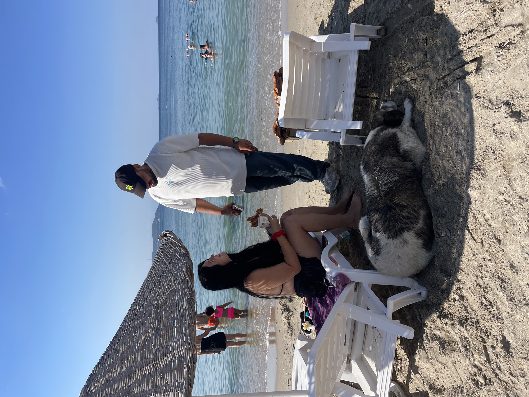 The image depicts a sunny day at a beach in Kadıköy, Turkey. In the foreground, a woman with long dark hair, wearing a black bikini top and black shorts, is seated on a white lounge chair under a woven umbrella. She is holding a cup and a piece of food, seemingly engaged in conversation with a man standing beside her. The man is wearing a white short-sleeved shirt, dark pants, and a baseball cap. A large, fluffy dog with brown and white fur is lying on the sand near the lounge chairs. In the background, the calm blue sea is visible, with several people swimming and enjoying the water. The shoreline is sandy, with other lounge chairs and a table laden with food. The overall atmosphere is relaxed and leisurely.