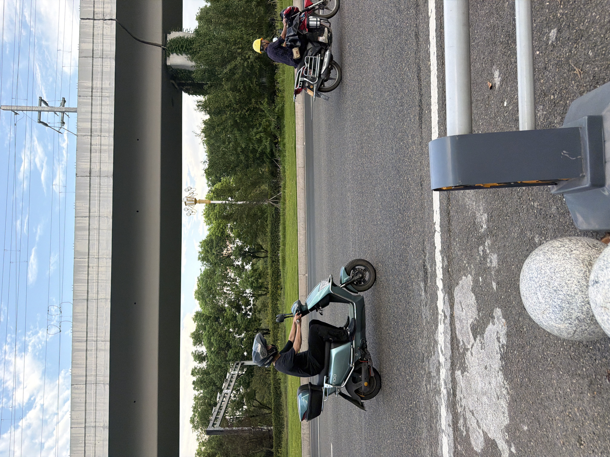 In Jiangjin, China, two individuals are seen riding motor vehicles on a paved road. In the foreground, a person wearing a black shirt and a black helmet is riding a teal electric scooter. The scooter has a black storage box mounted on the rear. Further down the road, a person wearing a dark blue shirt and a yellow helmet is riding a red motorcycle. The motorcycle has luggage strapped to the back and a chrome rack. An elevated overpass with visible power lines and concrete supports looms in the background, casting shadows on the road. The surrounding environment is characterized by lush green trees and a cloudy sky. In the immediate foreground, a grey metal barrier with rounded edges and two horizontal bars is positioned, and two speckled grey spheres are visible on the road's edge.