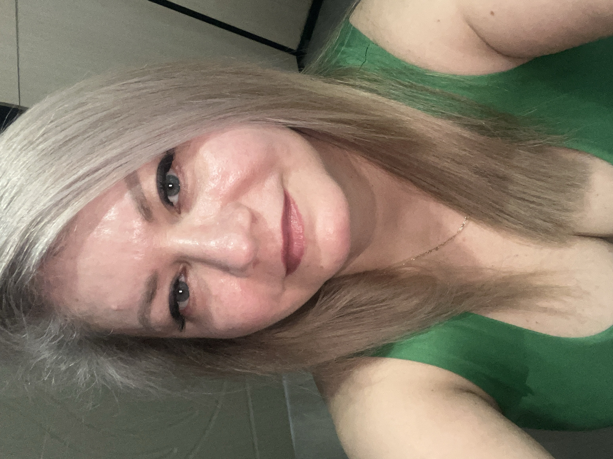 A selfie of a woman with blonde hair and blue eyes, wearing a green top. The location is not discernible.