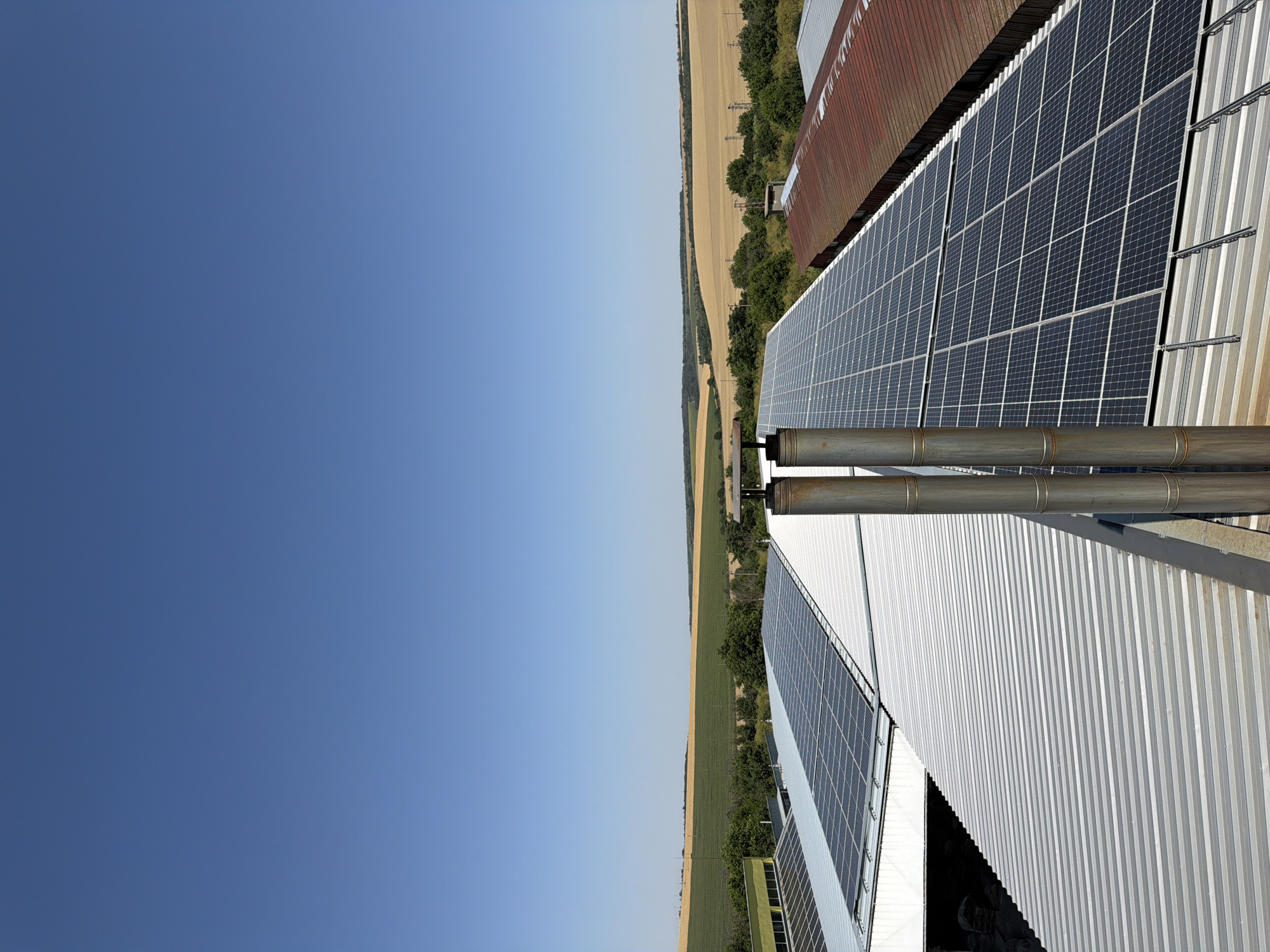 An elevated, eye-level shot captures a clear, bright blue sky above a rural landscape. In the foreground, the corrugated metal roof of industrial buildings dominates, covered with rows of dark solar panels. Two large, cylindrical metal smokestacks stand prominently on the roof. The buildings appear to be part of an industrial or agricultural complex. Beyond the buildings, a rolling landscape unfolds, featuring agricultural fields of varying colors, suggesting a mix of crops or stages of harvest, with scattered trees and utility poles dotting the horizon. The image was taken in Dolno Ablanovo, Bulgaria.