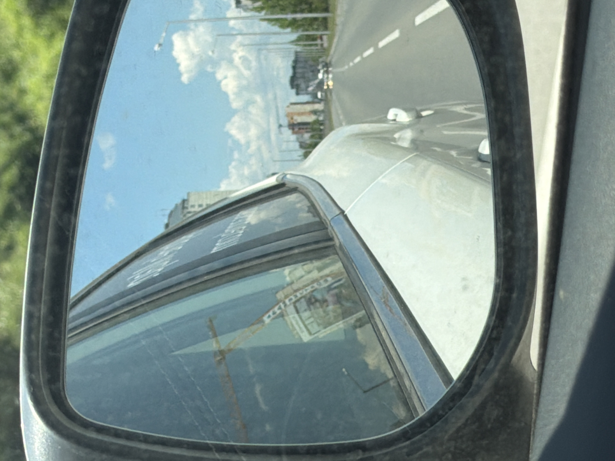 The image shows a reflection in a car's side mirror. The reflection shows a street scene with buildings and a bright blue sky with white clouds. In the foreground of the reflection, a yellow construction crane is visible. The car appears to be driving on a road with white lane markings. In the distance, several multi-story buildings are visible. The scene is consistent with an urban or suburban environment. There is no visible indication of the specific location within Sofia, Bulgaria, however the presence of construction and modern buildings suggests a developing area of the city.