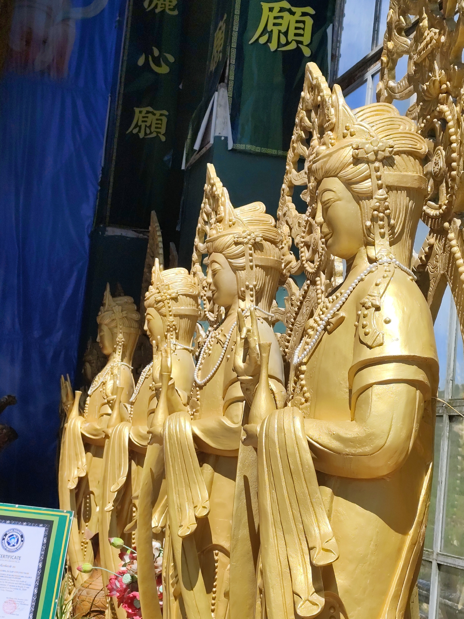 The image shows a line of five golden statues, likely religious figures, in what appears to be an outdoor setting. The statues are elaborately decorated with ornate headdresses, necklaces, and flowing robes. The figures are depicted from the waist up, with their heads slightly tilted and their hands held in various poses. In the background, a green banner with yellow lettering and a blue drape are visible. To the left of the statues, a certificate framed in green and white is partially visible, suggesting a place of significance or recognition. The statues are positioned in front of a large window with a grid pattern, revealing a blurry outdoor environment beyond. The overall impression is one of grandeur and religious devotion. The location is specified as Xuân Thành, Vietnam.