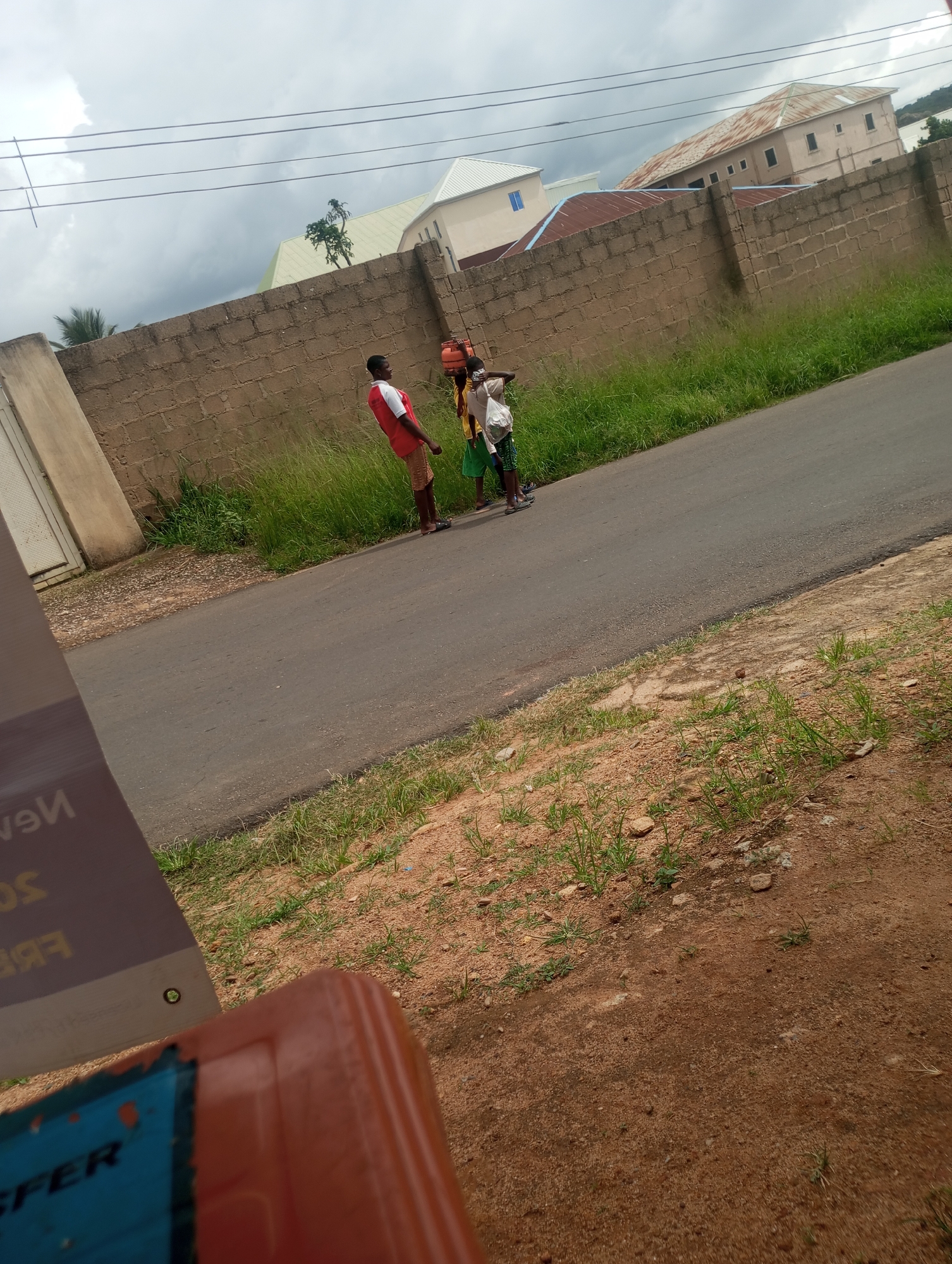The image depicts a cloudy daytime scene on a paved road in the city of Jos, Nigeria. Three young individuals are visible, standing near a tan brick wall. One person, wearing a red shirt and brown shorts, is facing away from the camera. Another, in a white and yellow outfit, is carrying an orange gas cylinder on their head. A third individual, wearing a green and black patterned skirt, is also present. The road is bordered by tall grass on one side and a red dirt embankment on the other. In the background, several buildings with corrugated iron roofs are visible behind the wall. The foreground shows a tilted orange object and part of a sign with blue and white lettering, partially obscuring the view.