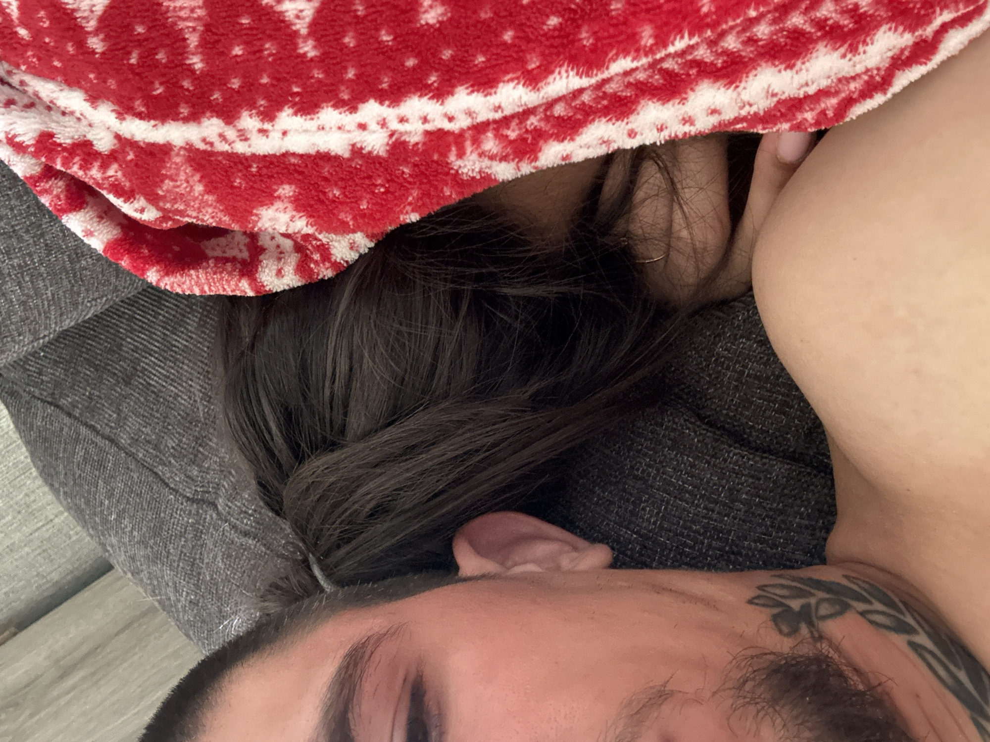 The image depicts a close-up, intimate scene indoors. A person with visible facial features including an eyebrow, eyelashes, and part of a forehead and ear is at the bottom of the frame. A tattoo is visible on their shoulder. Above and to the side of this person is another individual with long dark hair and a red and white patterned blanket partially covering them. The scene is situated on a gray upholstered surface, likely a sofa or bed. There is no information in the image that helps determine that the location of the image is in Gorna Malina, Bulgaria.