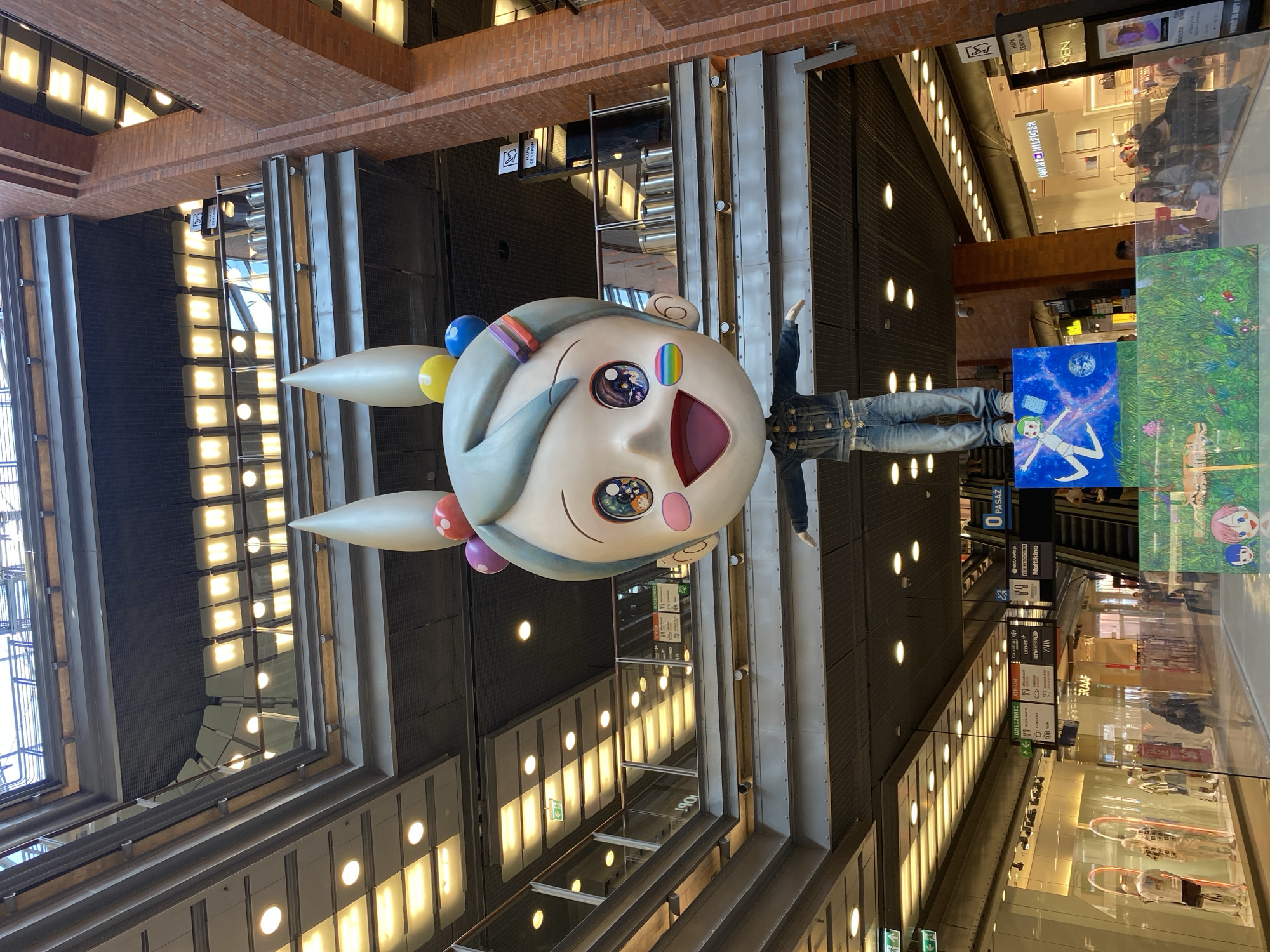 The image shows a large, whimsical sculpture of a character with an oversized head, big eyes, and ears, adorned with colorful balls and rainbow accents. The character is dressed in a denim jacket and jeans, with its arms outstretched. It stands on a platform that features two artistic paintings, one depicting a person floating in space and another showing a lush green field with stylized figures.
The sculpture is located in the Atrium of a shopping mall. The Atrium, a part of the mall, is characterized by its multi-level architecture with balconies, glass railings, and exposed metal beams. The lighting in the mall comes from rows of fluorescent lights and spotlights. Shops and escalators are visible in the background, indicating a commercial environment.
One of the signs visible in the mall reads "0 PASAŻ", which translates to "Passage 0". Another sign indicates "ATRIUM" and lists various shops and services, including "Carrefour", "RTV EURO AGD", and "Multikino". A sign for "TOMMY HILFIGER" is also visible on a shop front. The overall scene suggests an indoor shopping mall in a modern urban setting. The architectural style and the presence of shops such as "Tommy Hilfiger" are common in many European cities. Given the signage and the likely context of a prominent art installation in a mall, this scene is likely located in a major city. The Polish language on the signs strongly suggests the location is in Poland. Based on public information about notable art installations in shopping malls in Poland, this artwork is located in the **Posnania shopping mall in Poznan, Poland.**