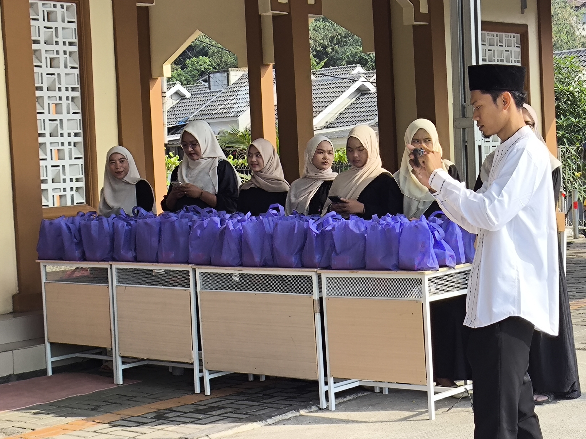 In Cilegon, Indonesia, a group of people are gathered outdoors, likely for an event or distribution. The scene is set in front of a building with tan and brown architectural elements and decorative white latticework windows. A long table is laden with numerous purple plastic bags, suggesting a distribution of goods or donations.
To the right of the table, a man wearing a white long-sleeved shirt, black pants, and a black peci (a cap) is holding a smartphone, appearing to take a picture or record a video. He is facing left, towards the table and the individuals standing behind it.
Behind the table, several individuals are present, predominantly women wearing hijabs. From left to right, a woman in a light-colored hijab is looking towards the viewer. Next to her, a woman in a similar hijab and glasses is looking down at her smartphone. A third woman, also in a light-colored hijab, is looking to her left. To her right, another woman in a light-colored hijab is looking down at her smartphone. Further to the right, a woman in a dark hijab and an additional person in a dark outfit are partially visible. The background reveals rooftops and trees, indicating a residential or suburban area. The ground is paved with bricks and features some painted lines.