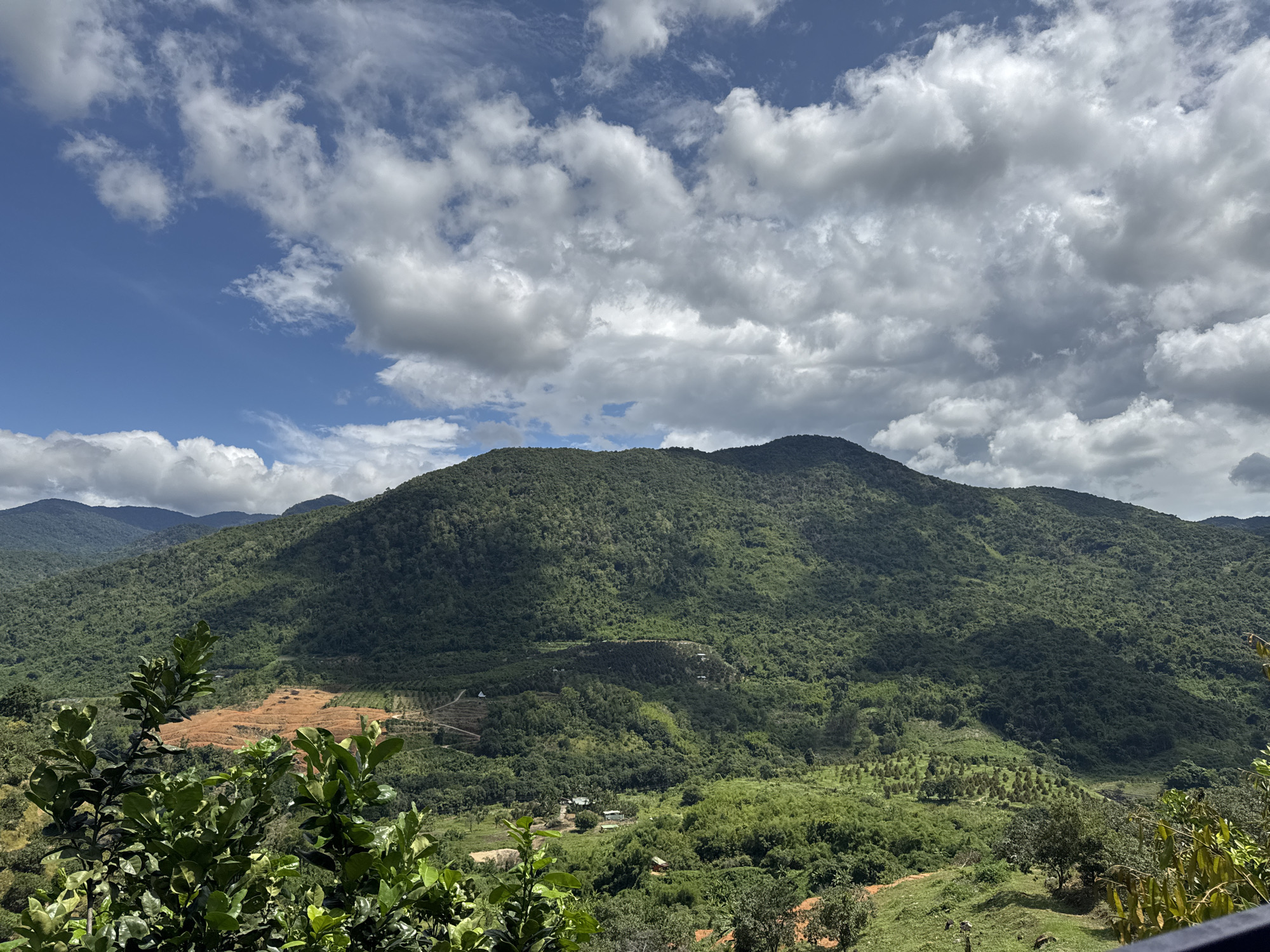 The image captures a scenic vista of rolling green mountains under a partially cloudy blue sky. In the foreground, lush green foliage is visible, framing the expansive landscape. The midground features a cleared, reddish-brown patch of land on a hillside, likely for agricultural or development purposes, contrasting with the dense, verdant vegetation that covers the rest of the terrain. Scattered amidst the greenery, several small buildings and structures can be observed, suggesting the presence of a rural settlement. The background is dominated by layers of mountains receding into the distance, their slopes uniformly covered in thick vegetation. The scene depicts a natural environment with evidence of human presence in the form of cleared land and buildings. The location is identified as La Dạ, Vietnam.