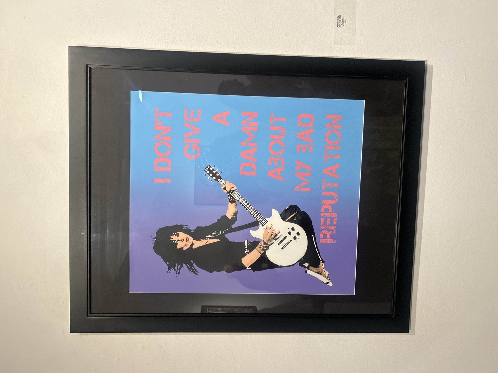 A framed poster of Joan Jett playing guitar and featuring the phrase "I DON'T GIVE A DAMN ABOUT MY BAD REPUTATION" hangs on a white wall. A label with text is affixed above the frame. The scene is indoors. The location is unknown.
