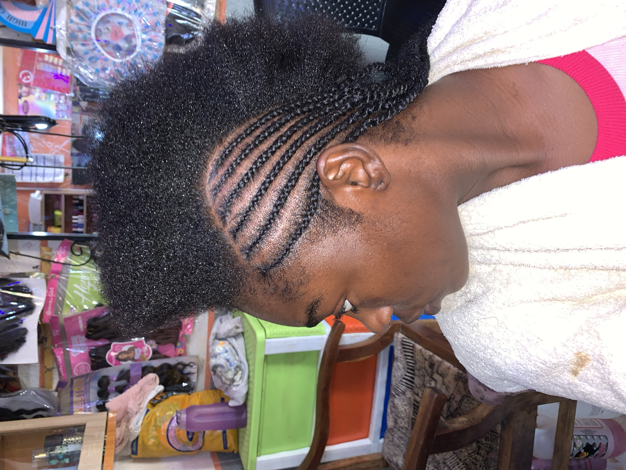 A woman with cornrow braids at the temple and afro-textured hair on top is sitting indoors. She is wearing a white towel and a red garment. The background shows hair care products, including packs of hair extensions and shower caps. The location is Rafin Par, Nigeria.