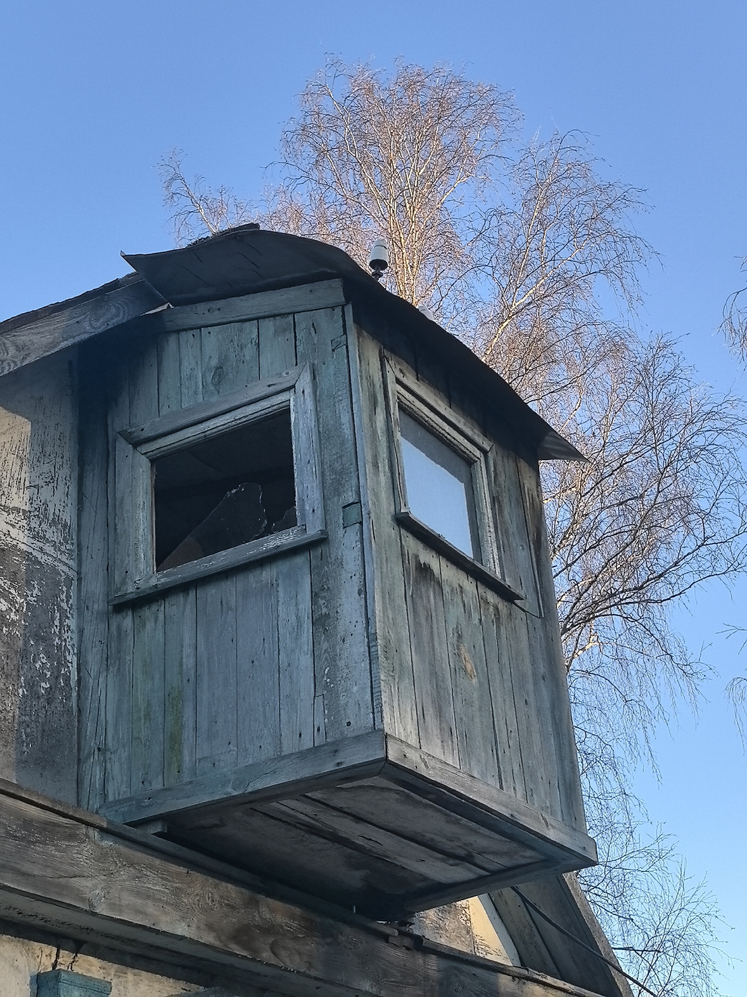 An exterior shot depicts a weathered wooden structure with two windows, extending from the side of a building. One window is boarded up, while the other is open. Above, there is a peaked roof. The location is likely Noviy, Russia. A leafless tree is in the background against a blue sky.