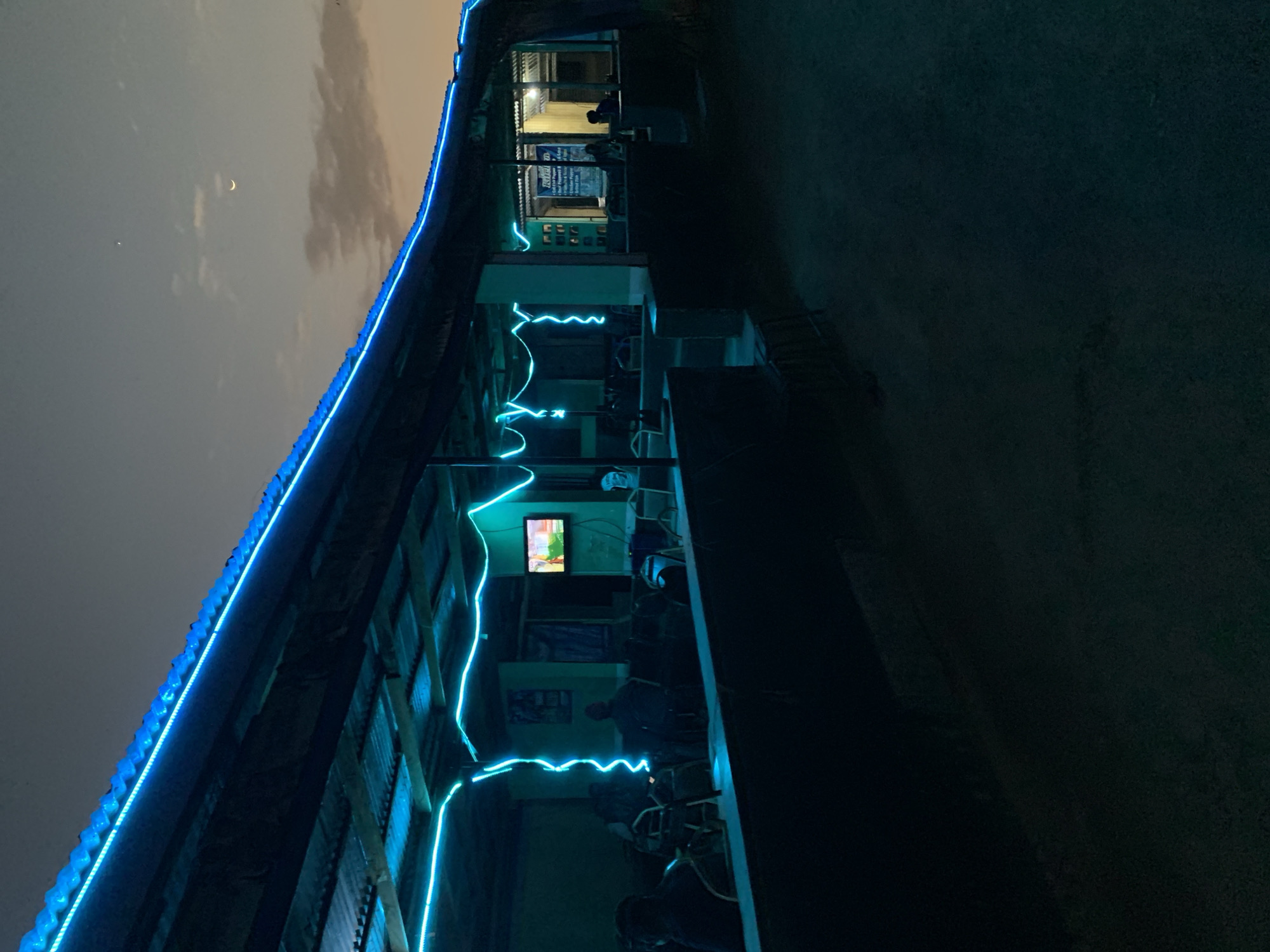 The image depicts a two-story building in New Stadium, Nigeria, at night. The building is adorned with bright blue LED lights tracing the roofline and zigzagging along the facade. A crescent moon is visible in the night sky. People are present inside the building; one is seen in the upper level. A television screen is illuminated on the second floor. Outside the building is a dark, paved area.