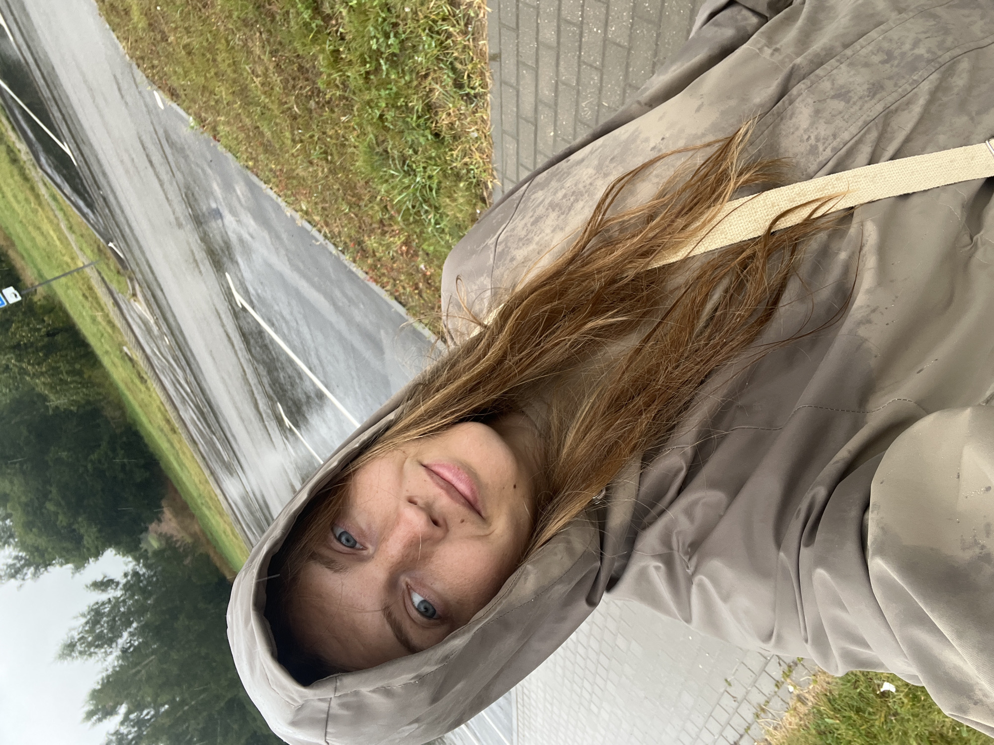 In an outdoor, overcast scene in Сеніцкі сельскі Савет, Belarus, a young woman with long, auburn hair and blue eyes, wearing a grey hooded jacket, poses for a selfie. The jacket appears wet, as does the surrounding environment. She stands on a paved walkway adjacent to a grassy area, with a wet asphalt parking lot visible behind her, marked with white lines. Trees form a backdrop. The weather is clearly rainy.