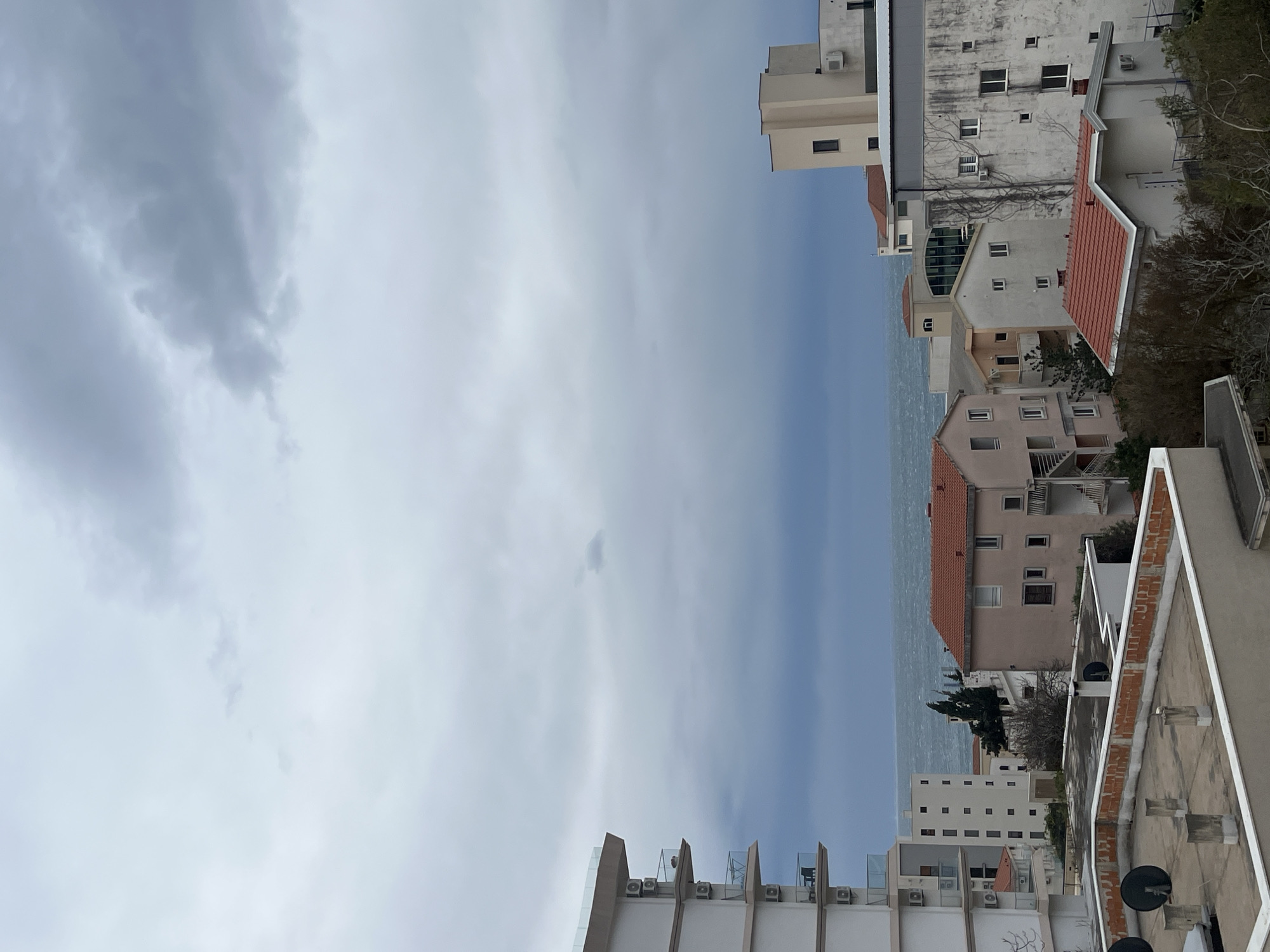 The image shows a view over buildings towards the sea in Boreti, Montenegro. The sky is overcast with heavy grey clouds. The sea appears calm, with a band of blue-grey water visible between the buildings. The foreground is dominated by the rooftops of several buildings, varying in height and color. There are a mix of residential and commercial buildings. Many buildings have air conditioning units on the exterior. The overall impression is one of a densely built urban environment with a coastal location.