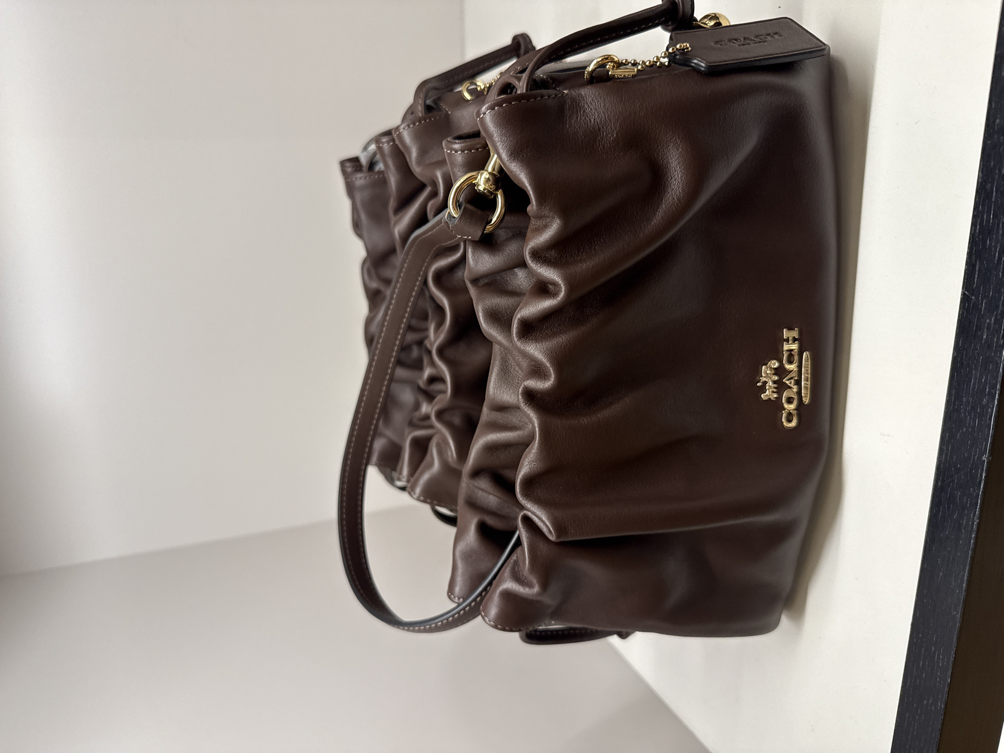 The image shows a brown Coach purse with a gathered or ruched design. The purse is positioned against a white surface, likely a shelf or wall, with a dark-colored trim or frame visible to the right. The purse features a gold Coach logo and hardware. The location is in the city of Sunrise, United States of America.