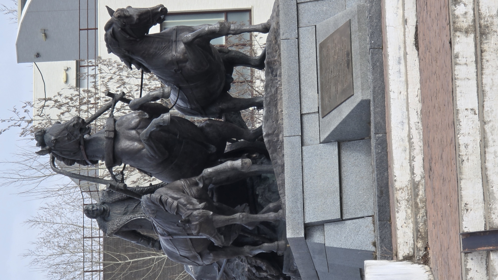 The image captures a bronze monument in Lensk, Russia. The monument features a dynamic composition of horses and riders, appearing to charge forward. The statue is mounted on a gray stone base, and a bronze plaque is affixed to one of the stone blocks. In the background, there is a bare tree and a modern building with windows. Part of a damaged wooden structure is to the right.