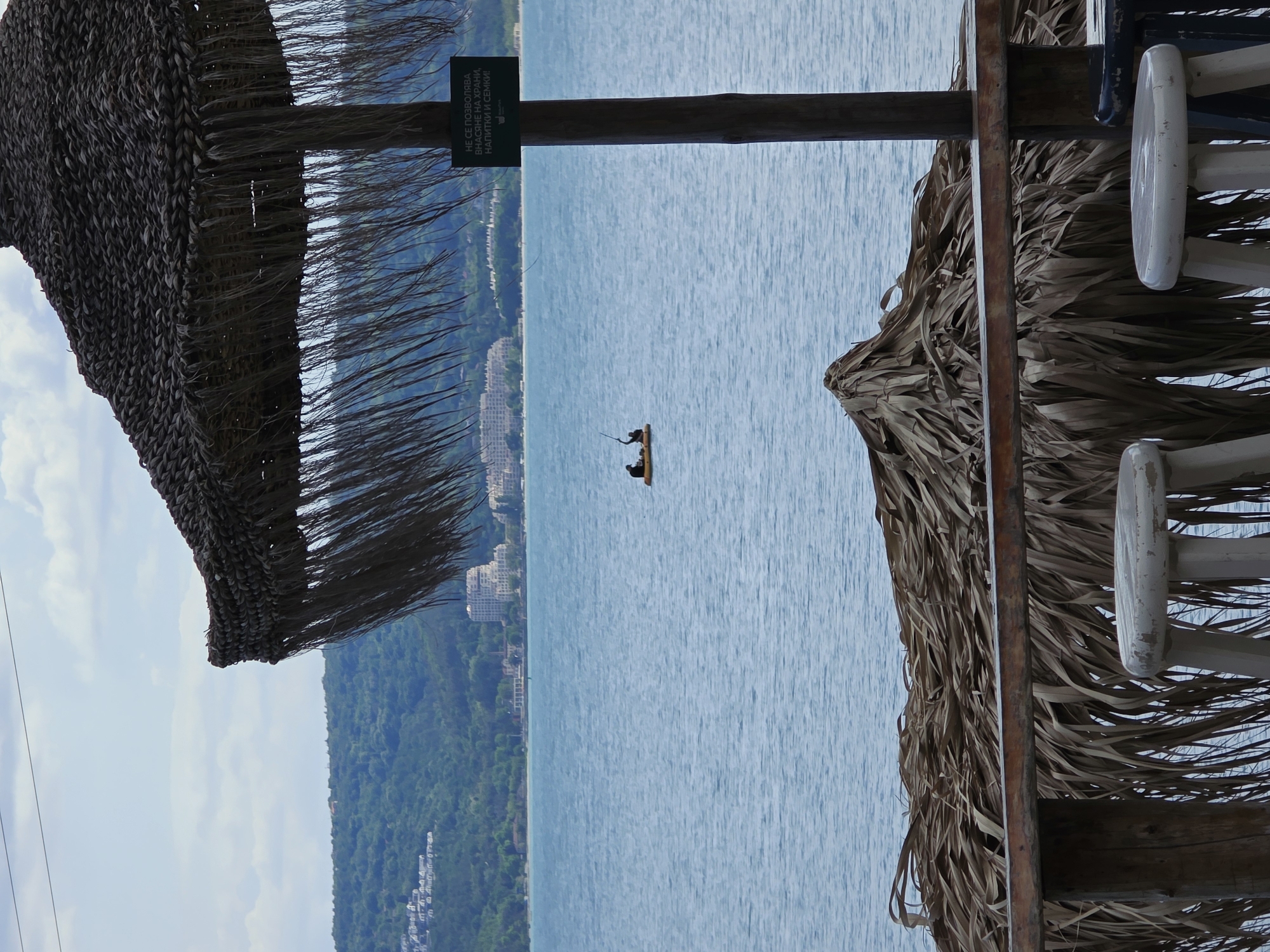 The image, taken in Varna, Bulgaria, shows a tranquil sea view framed by two thatched umbrellas. In the distance, two figures are in a boat, engaging in fishing, as suggested by the presence of a fishing rod. The backdrop reveals a coastal city with buildings and green forests, creating a scenic contrast. The umbrellas, constructed with wooden frames, add a rustic touch to the scene. A sign is seen with indiscernible writing.