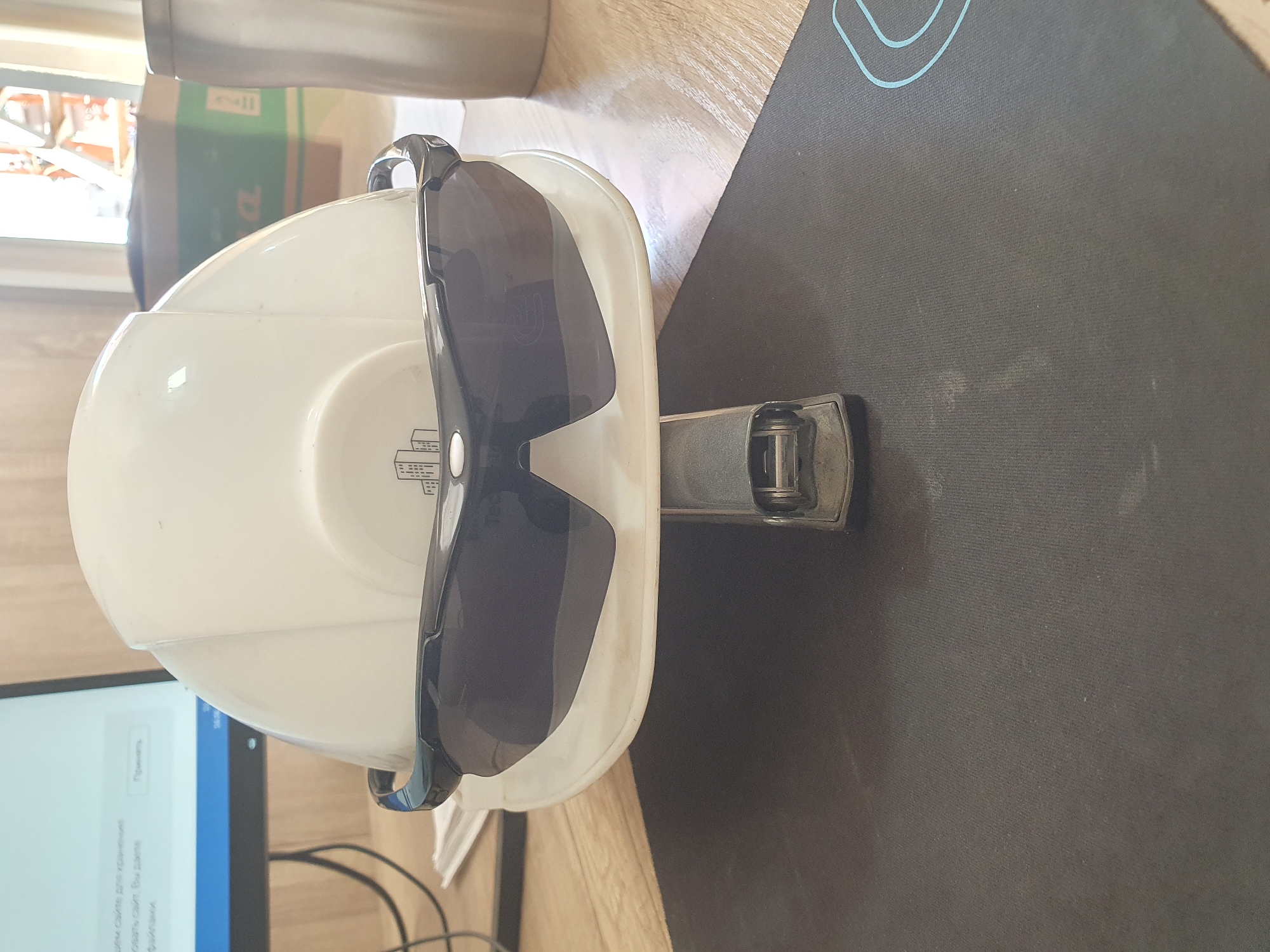 A white construction hard hat, adorned with black sunglasses, is positioned upside down on a silver stapler. The assembly rests on a black mousepad with a light blue stylized pattern. In the background, a computer monitor displays a website interface with Russian text. To the right, a metal thermos and a green cardboard box are visible. The scene is illuminated by natural light, suggesting an indoor environment, possibly an office or workshop, in Tashkent, Uzbekistan. There are no people or animals present.