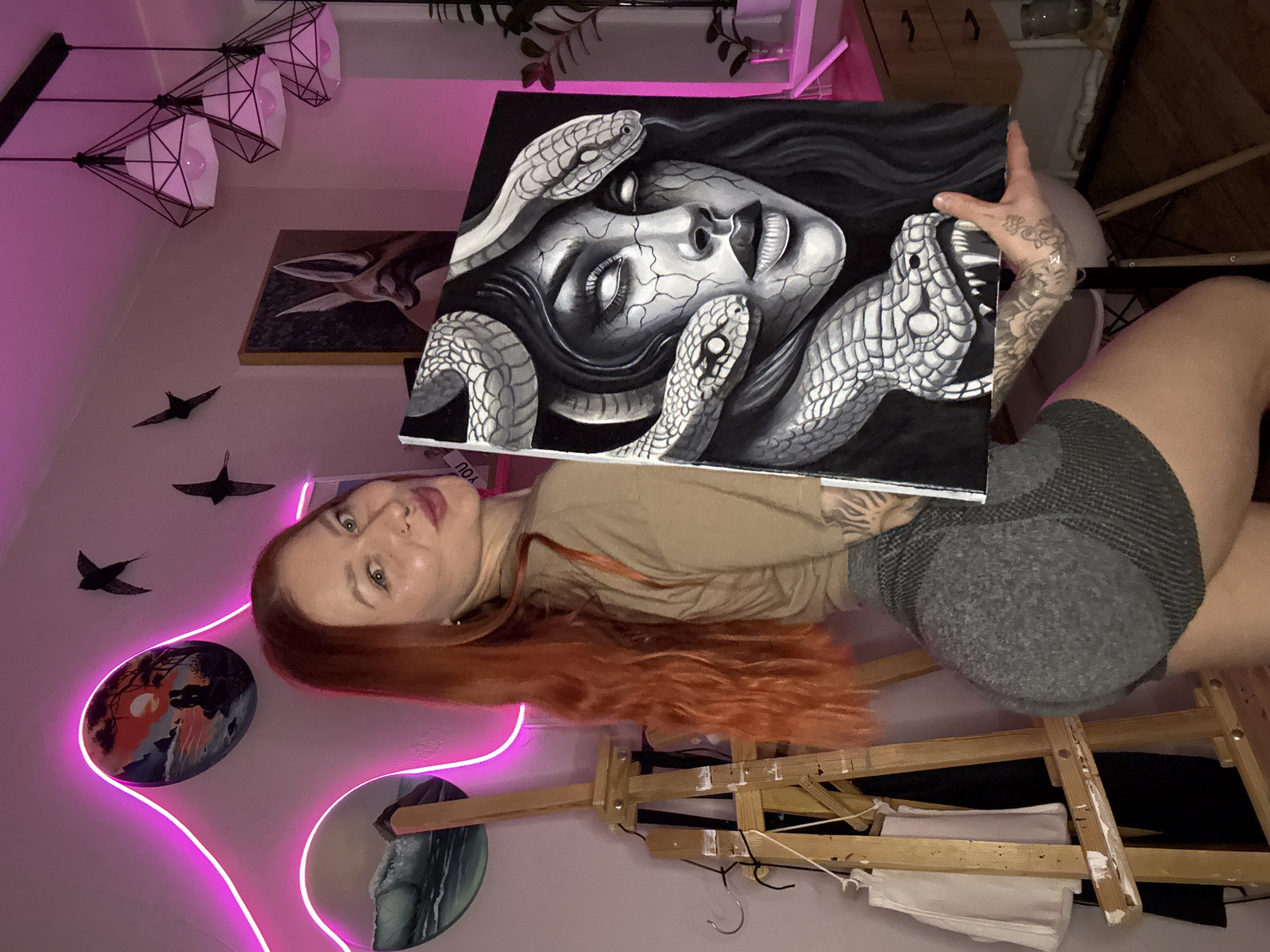 A red-haired woman poses in front of a wall with decorative elements in an indoor space, possibly in Rostov-on-Don, Russia. She holds a painting of a grayscale Medusa head surrounded by snakes. The room features modern lighting fixtures, artistic wall decorations, and an easel. The woman has tattoos on her arm. She wears gray shorts and a beige top.