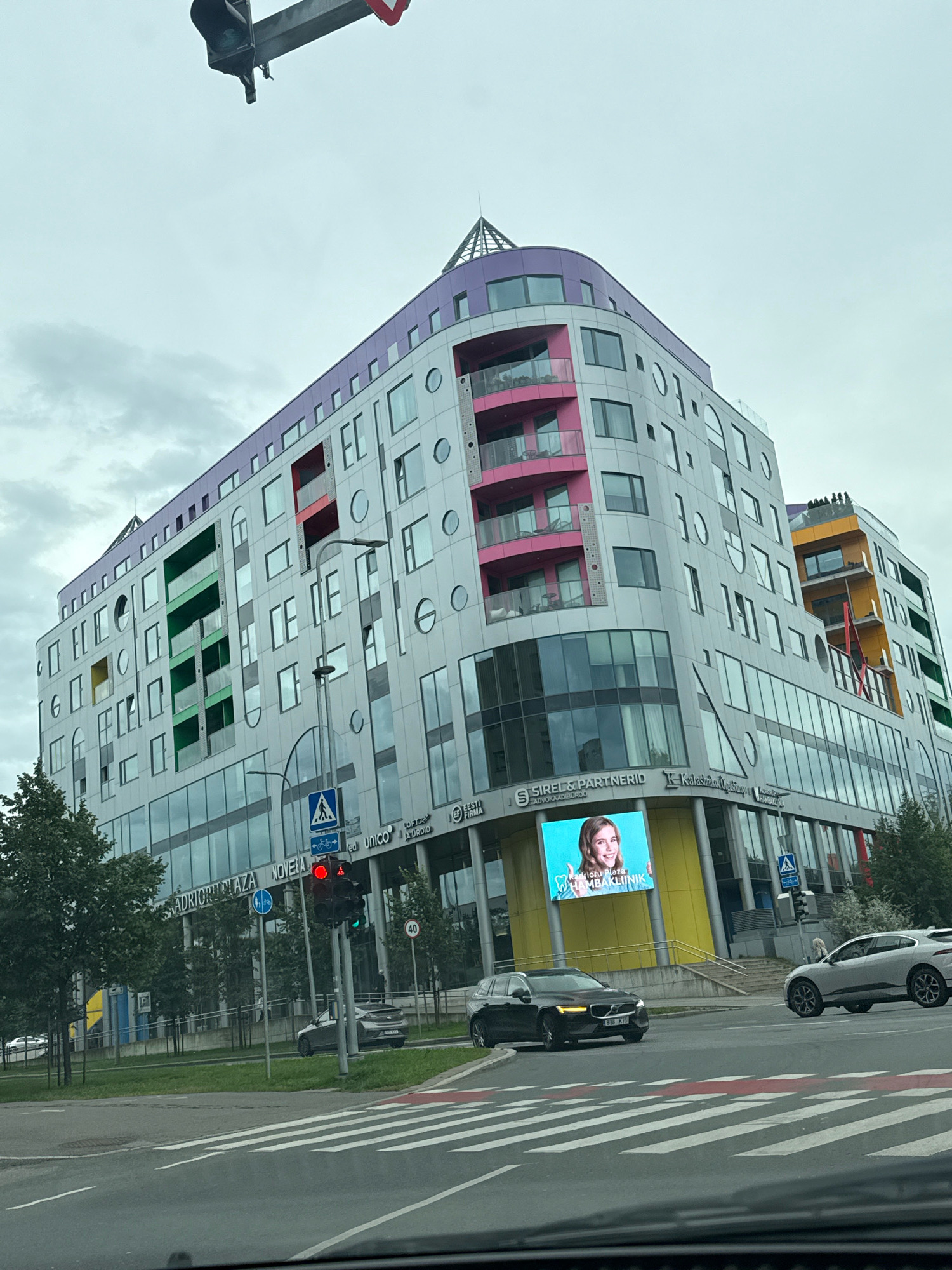 The image shows a street scene in Tallinn, Estonia, focusing on a modern, multi-colored building with a distinctive architectural design. The building features a curved facade with sections of grey, pink, green, and purple. It has numerous windows of various shapes and sizes, including circular ones. On the ground floor, there are large glass windows with several businesses advertised, such as "SIREL & PARTNERID" and "HAMBAKLINIK". A large digital billboard on the building displays a smiling woman.
In the foreground, a street with a pedestrian crossing is visible. A traffic light is illuminated red, indicating a stop. Two cars are present: a dark-colored Volvo station wagon in the left lane and a silver Jaguar SUV in the right lane, both appearing to be at or approaching the intersection. A traffic sign with a bicycle symbol is also visible. Trees line the sidewalk in front of the building. The sky is overcast. The overall impression is an urban environment with contemporary architecture and active traffic.
