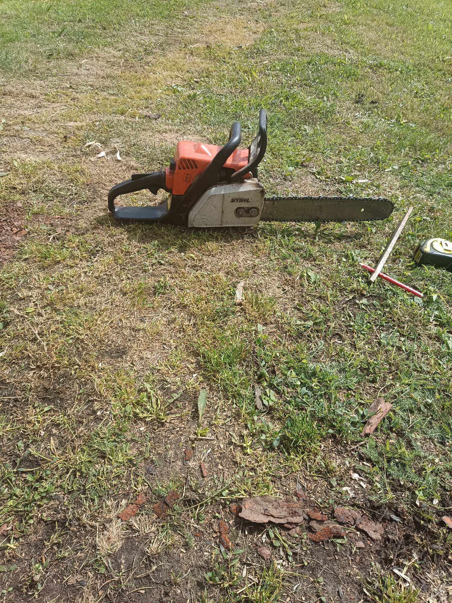A Stihl chainsaw lies on its side on a grassy area, with its chain bar facing to the right. The chainsaw is orange and silver. To the right of the chainsaw, a yellow and black measuring tape, a red pencil, and a wooden ruler are placed on the ground. The ground is covered in dry grass and patches of green grass, with some scattered dirt and small pieces of bark. The image appears to be taken outdoors on a sunny day, likely in a yard or park within Удзелаўскі сельскі Савет, Belarus.