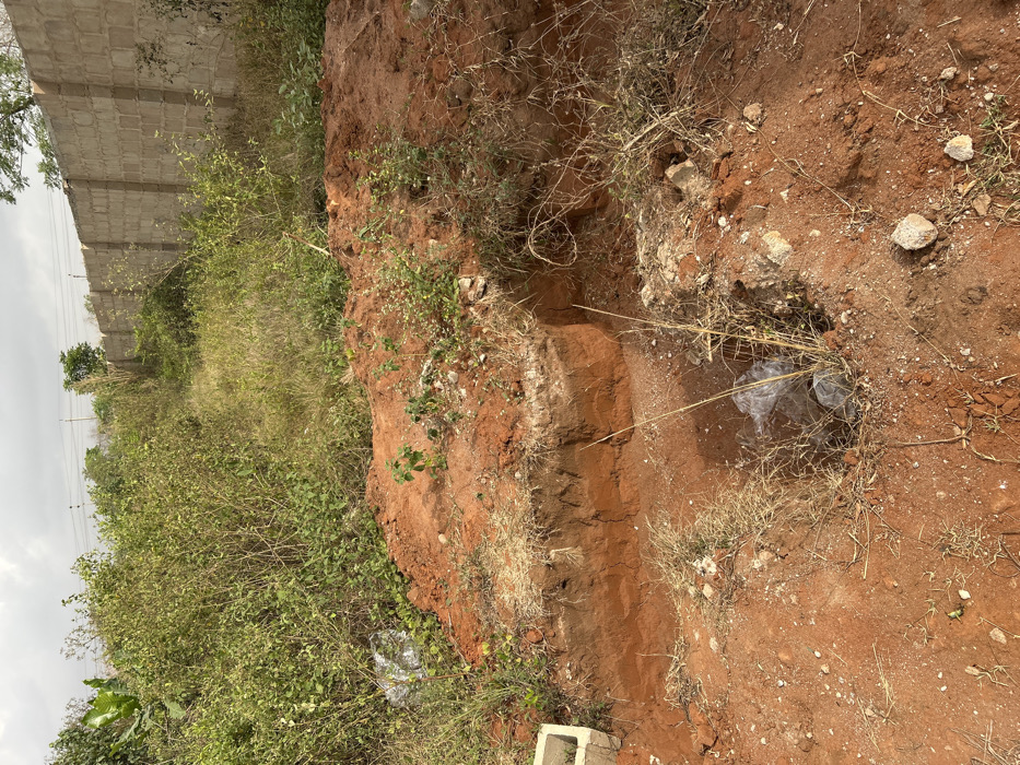 The image displays an outdoor scene in Ilishan, Nigeria, characterized by disturbed reddish-brown earth and dense vegetation under an overcast sky. In the immediate foreground, a roughly rectangular trench, approximately 1-2 feet deep, is excavated into the soil, revealing distinct layers. To the left of this trench, a partial concrete block is visible. Directly in front of the trench, a smaller, irregular circular pit contains translucent plastic debris. The ground surface features scattered dry grass and small rocks. Behind the main trench, a mound of freshly excavated reddish-brown soil is present, with small green plant sprouts growing on it. The midground and left background are densely overgrown with unkempt green bushes and tall grass. In the right background, a tall, light-colored concrete block wall, topped with what appears to be barbed wire or an electric fence, runs horizontally. Faint utility lines are discernible above the vegetation in the far background. No individuals or specific activities are observed.