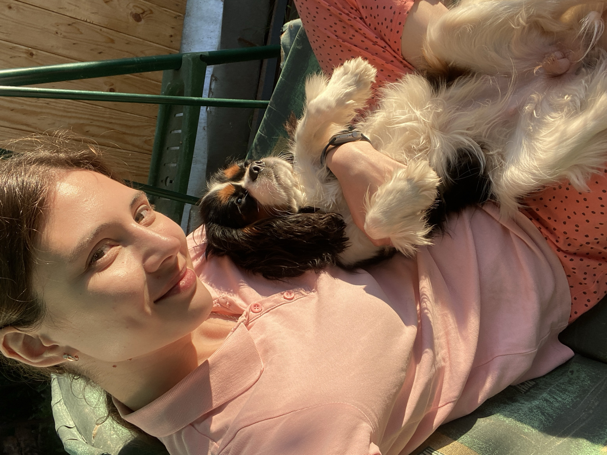 A young woman with brown hair is sitting outdoors, holding a small dog. She is wearing a pink polo shirt and pink polka dot pants. The dog, a Cavalier King Charles Spaniel, has black, white, and tan coloring. They are seated on a green chair, likely in a backyard or patio area, with a wooden fence visible in the background. The scene is illuminated by natural sunlight, casting warm shadows. The woman is looking towards the camera with a slight smile, while the dog appears relaxed, with its eyes closed. The overall setting suggests a casual, outdoor leisure activity.