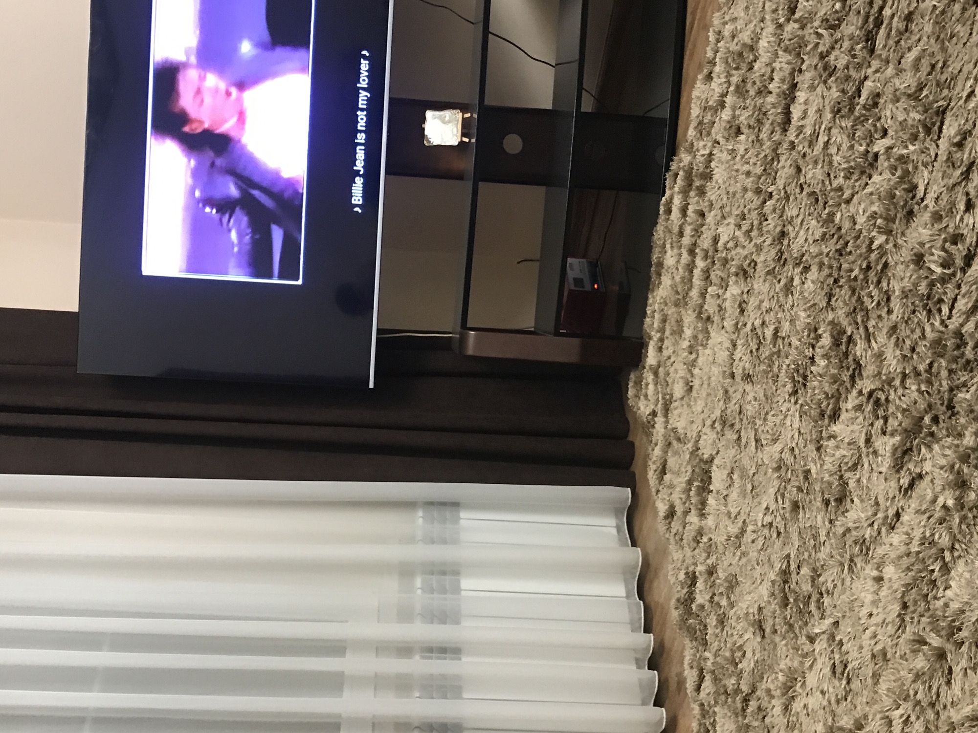 The image depicts a room interior. A television screen displays a video with the words "Billie Jean is not my lover." Below the TV is a set of shelves with objects on them. A shaggy rug covers part of the floor. A window with white blinds is also visible. There is no information to ascertain its location is in the city of Yerevan, Armenia.