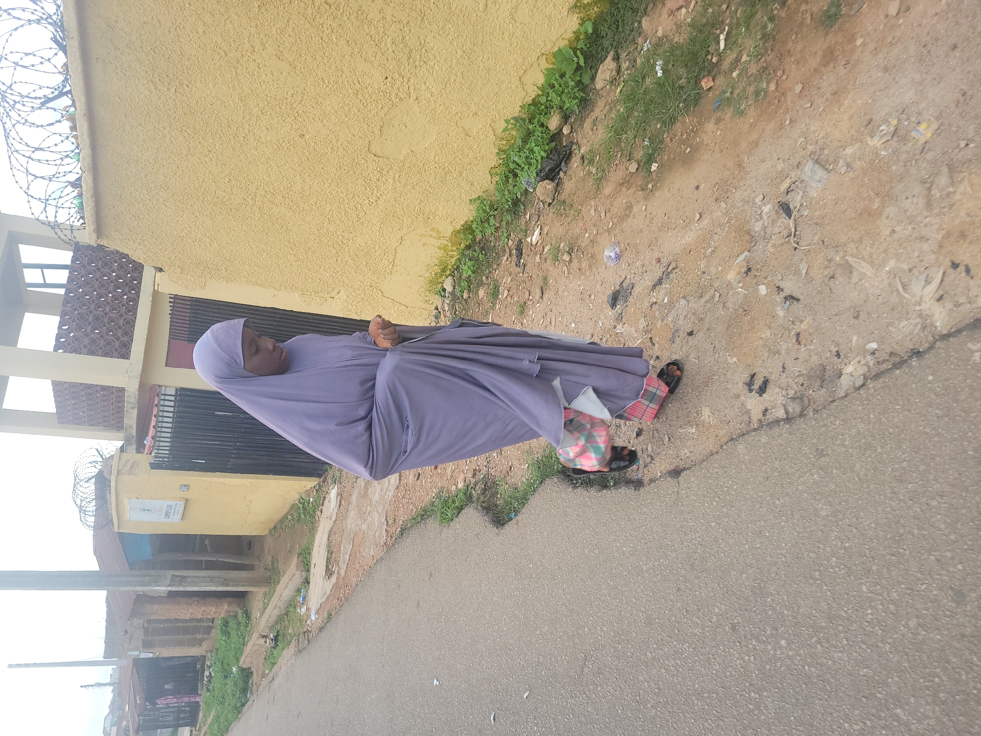 A young female, wearing a light purple hijab and a matching loose-fitting garment, walks along a dusty, paved road. Her feet are clad in dark sandals with colorful patterned straps. She is looking downwards, with her hands clasped together near her waist, appearing to be lost in thought or perhaps praying. The road is bordered by a strip of dirt and sparse green vegetation, with some scattered debris and plastic waste. To her right, a tall, textured yellow wall dominates the background, topped with a coiled wire fence. Behind the wall, a building with a light-colored facade and a dark patterned element is visible. To her left, a more modest collection of buildings with corrugated metal roofs and brick walls line the road, suggesting a more informal or residential area. The scene is likely an urban or semi-urban environment in Jos, Nigeria, characterized by its distinct architecture and the blend of paved and unpaved surfaces.