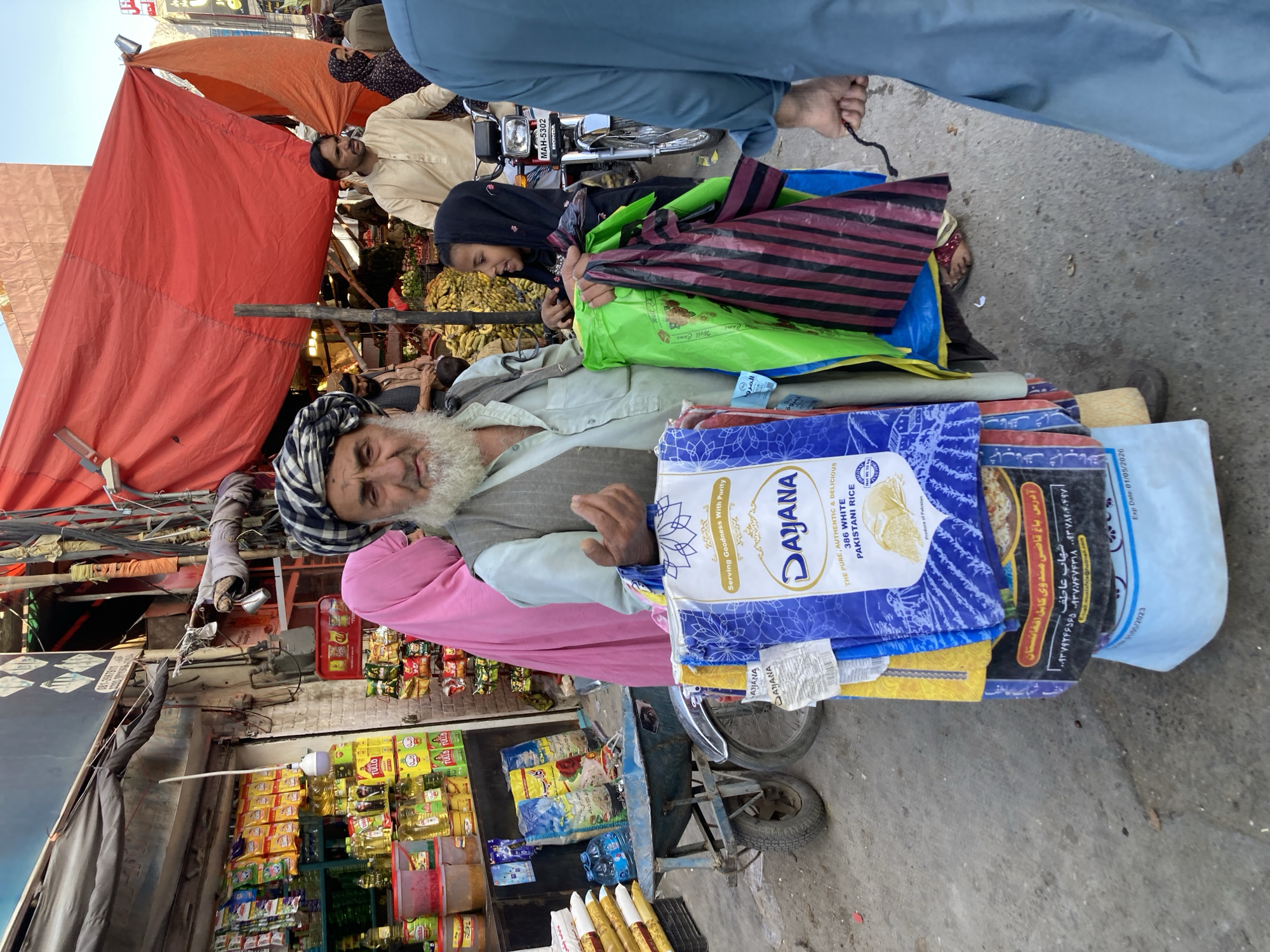 This image captures a vibrant, bustling street market scene, likely during daytime under clear weather, within Quetta City Tehsil, Pakistan. The perspective is slightly elevated, looking down into the activity.
At the center, an elderly man with a long white beard and a distinctive black and white checkered turban is prominent. He wears a light-colored long-sleeved shirt and a pink shawl draped over his shoulder, and appears to be pushing a two-wheeled handcart laden with goods. On the cart, multiple large bags are visible, including blue and white bags of "DAJANA 386 WHITE PAKISTANI RICE," advertised as "Serving Goodness With Purity." Another bag displays an address in "Bagh-e Qazi Mandawi Kabul, Afghanistan," along with Afghan phone numbers for "Shahab Atif," and an expiry date of "01/05/2026," suggesting cross-border trade or influences common in the region.
To the right of the elderly man, a young girl with dark hair covered by a dark headscarf is seen holding several colorful shopping bags, including a bright green one and a striped maroon and black one. Further right, a man in a light-colored shalwar kameez stands near a motorbike with the license plate "MAH-5302." A person in a light blue outfit is partially visible at the top, their hand holding what appears to be a black string.
The background reveals a lively market environment with makeshift stalls covered by red and orange tarpaulins. Various shops line the street, displaying packaged food items, bottles of oil, and other goods. On the upper left, utility wires are tangled overhead. The overall scene depicts a busy commercial thoroughfare.