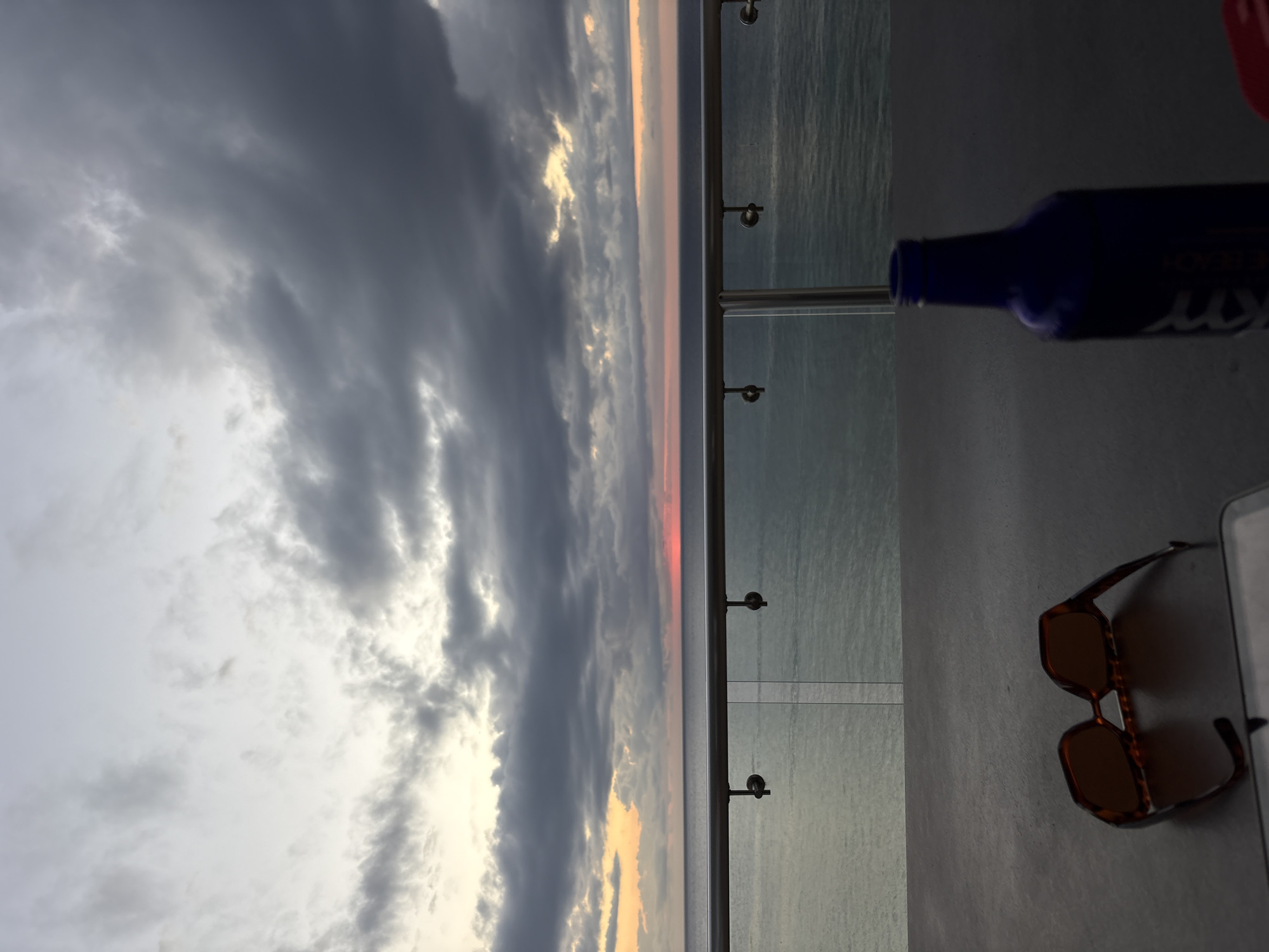 The image captures a view from what appears to be a balcony. The left side of the image displays a sky filled with grey and white clouds. There's a hint of red and yellow sunlight peeking through. A glass railing with metal supports separates the sky and sea from a grey, flat surface. On this surface, there's a dark blue bottle laying on its side, and a pair of brown tortoise-shell sunglasses. A white object is also visible beside the sunglasses. The location is likely in San Miguel de Cozumel, Mexico, as the presence of the ocean would indicate.