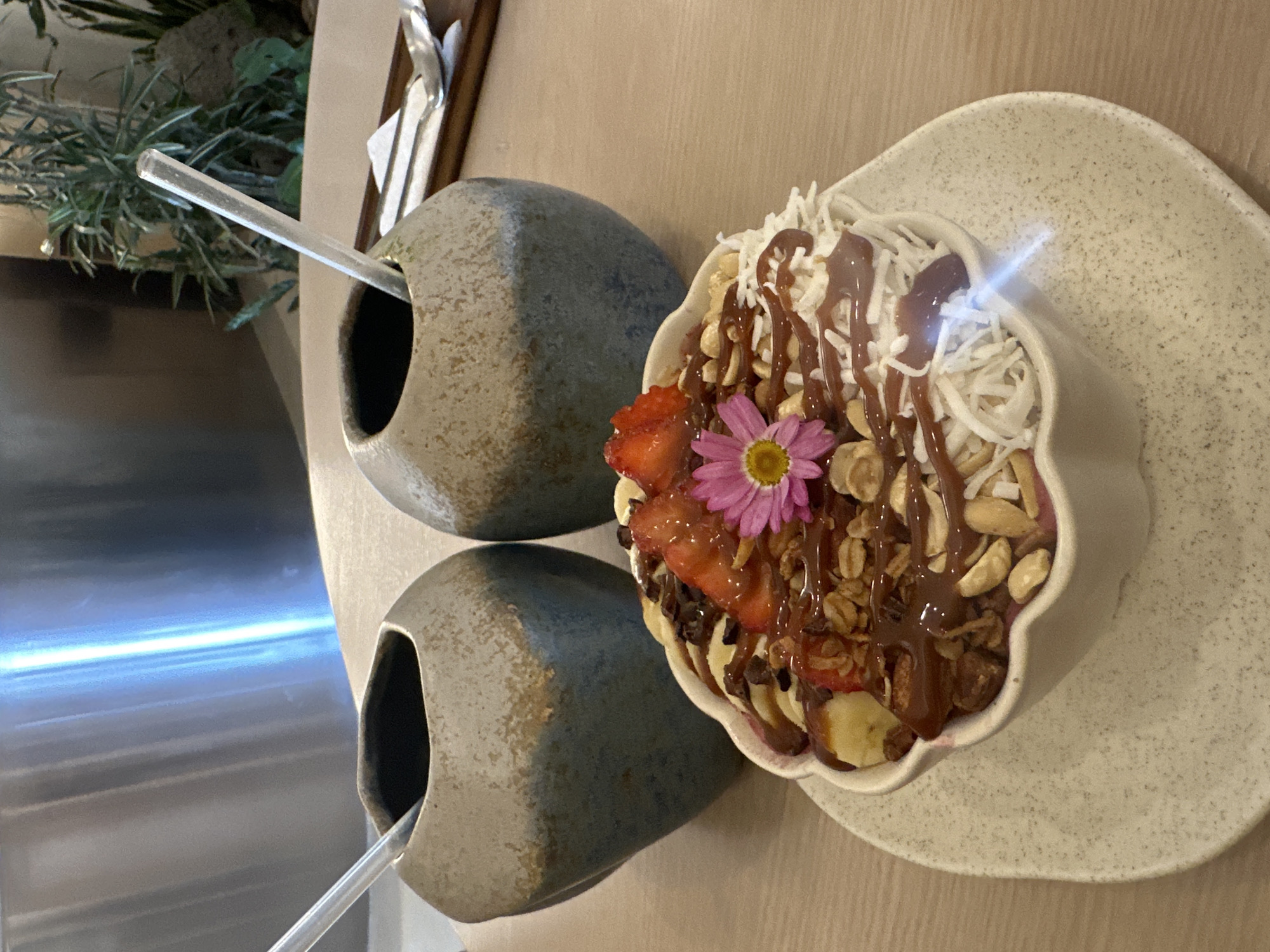 Here is a description of the image:
The image shows a close-up of a meal, possibly a smoothie bowl, on a table. The bowl contains fruit, granola, nuts, coconut shavings, and a drizzle of caramel or chocolate sauce. A pink flower is placed on top as decoration. The bowl sits on a speckled, light-colored plate.
Next to the bowl are two small, gray, stone-like containers, each with a clear straw or spoon sticking out. These containers may hold additional toppings or liquids.
The setting appears to be a restaurant or cafe. There are plants visible in the background. The image might be taken in Puebla, Mexico, given the prompt's mention of the city. The table's surface is a light-colored wood. The metal equipment in the background reflects the blue sunlight.