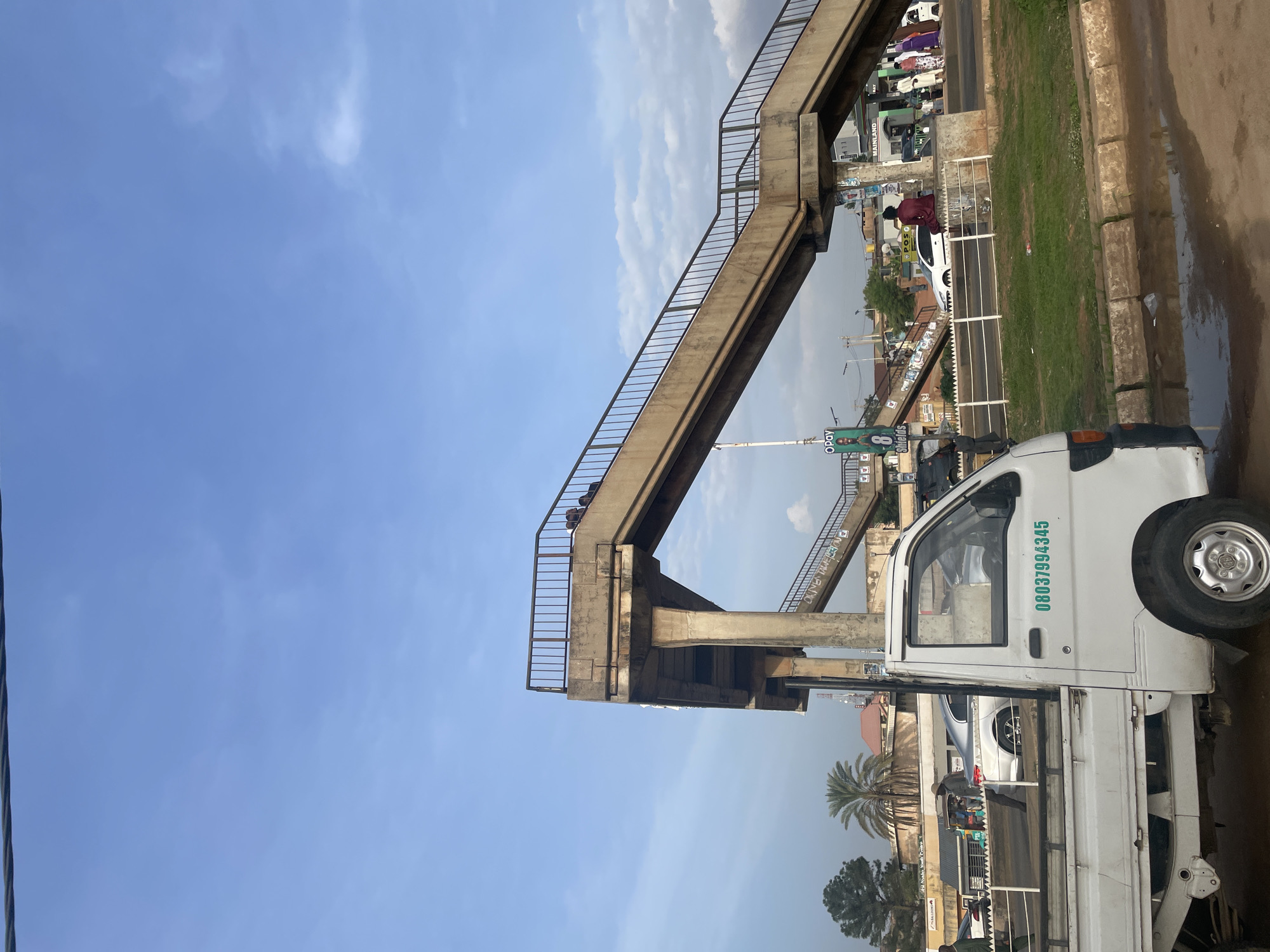 The image depicts a street scene in Gura Top, Nigeria. A white pickup truck is parked in the foreground. Above the truck, a concrete pedestrian overpass spans the road, with people visible on it. In the background are buildings, trees, and other vehicles. The sky is blue with scattered clouds.