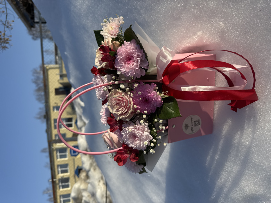 A pink floral arrangement, presented in a pink paper gift bag, is positioned on a field of white snow under a clear blue sky in Chaikovsky, Russia. The arrangement features light pink roses, various shades of pink and purple chrysanthemums, dark red alstroemeria, and white baby's breath, interspersed with green leaves. The pink gift bag includes two curved pink wire handles and has a red ribbon tied around its front, with a white ribbon partially visible beneath it, displaying "LIA". A circular white sticker on the bag's front bears the Russian text "ЛЕТО цветы и подарки" (SUMMER flowers and gifts) and a QR code. In the midground, a significant snowdrift rises behind the floral display. Beyond the snowdrift, a yellow, single-story building with numerous windows is visible, suggesting a public or commercial structure. Sparse, bare trees are discernible in the distant background. The scene is brightly illuminated by direct sunlight, casting distinct shadows across the snow. No people or animals are present.