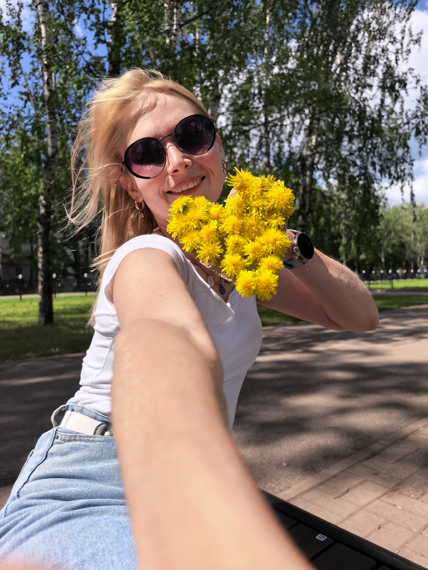A young blonde woman wearing sunglasses, a white t-shirt, and denim shorts smiles at the camera as she holds a bouquet of yellow dandelions. She is outdoors in a park with trees and a paved path in the background. The woman is in the foreground of the image, and her arm is extended towards the camera as if taking a selfie. She is wearing a watch on her left wrist. The overall scene appears to be a sunny day in a park.