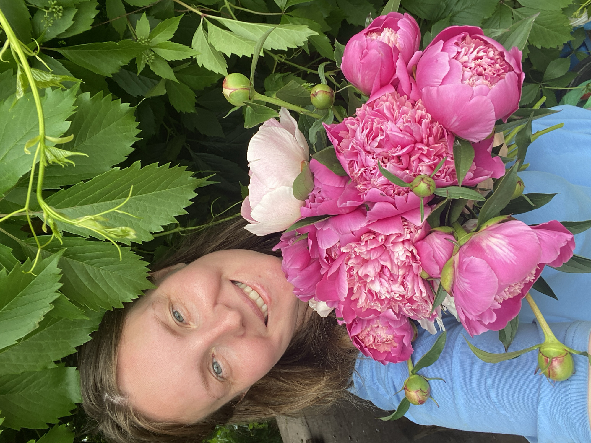 A woman with light brown hair and blue eyes is holding a bouquet of pink and white peonies. She is wearing a blue t-shirt and is partially obscured by green leaves. The background consists of dense foliage, suggesting an outdoor setting, possibly a garden or park. The image was taken in Chaikovsky, Russia.