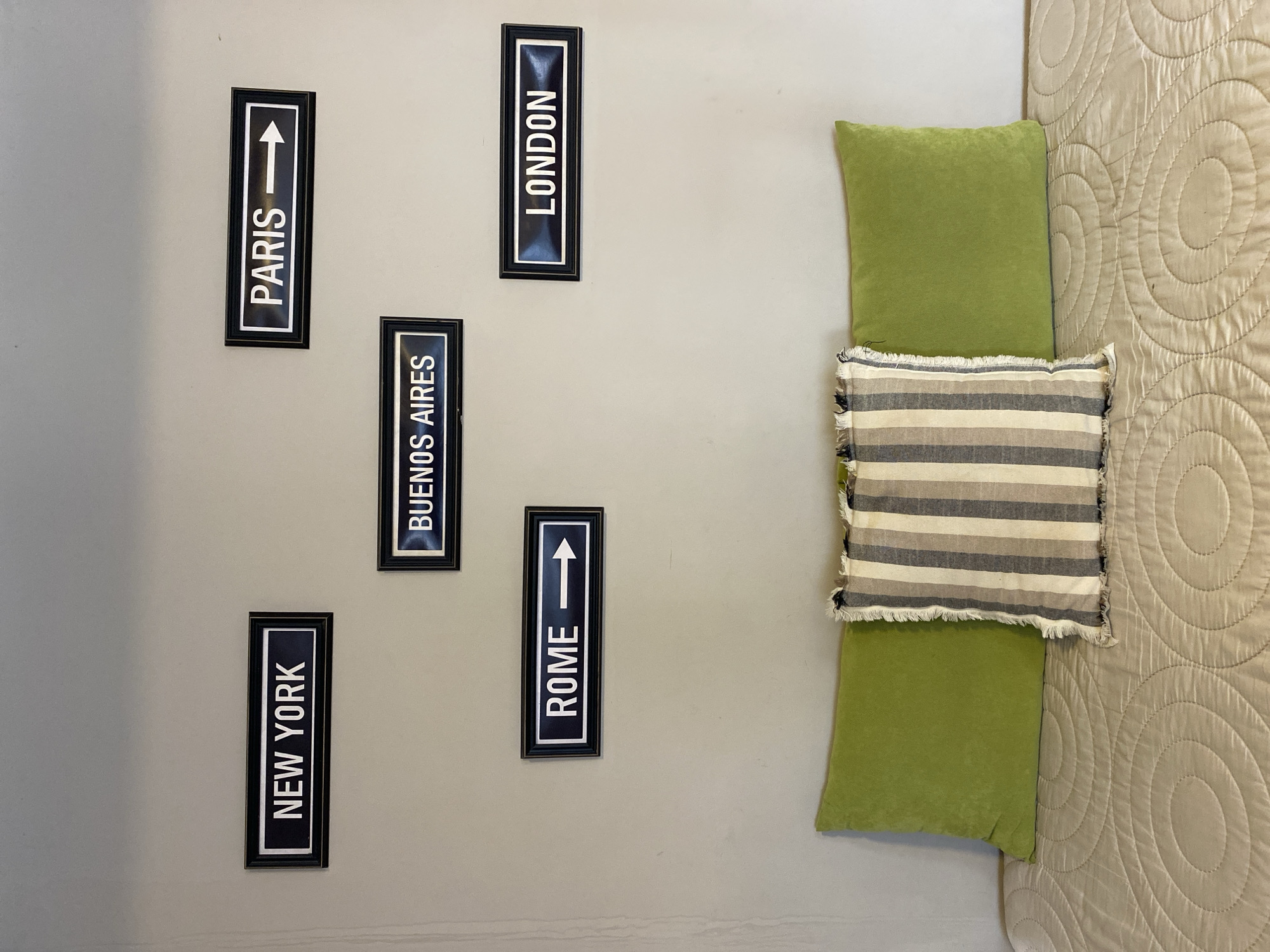 The image shows a wall with five framed signs indicating directions to various cities: Paris, London, Buenos Aires, Rome, and New York. To the right of the wall is a bed with a green pillow and a striped pillow on top, covered with a beige patterned bedspread. The location is in Buenos Aires, Argentina, indicated by the framed sign with that city's name.