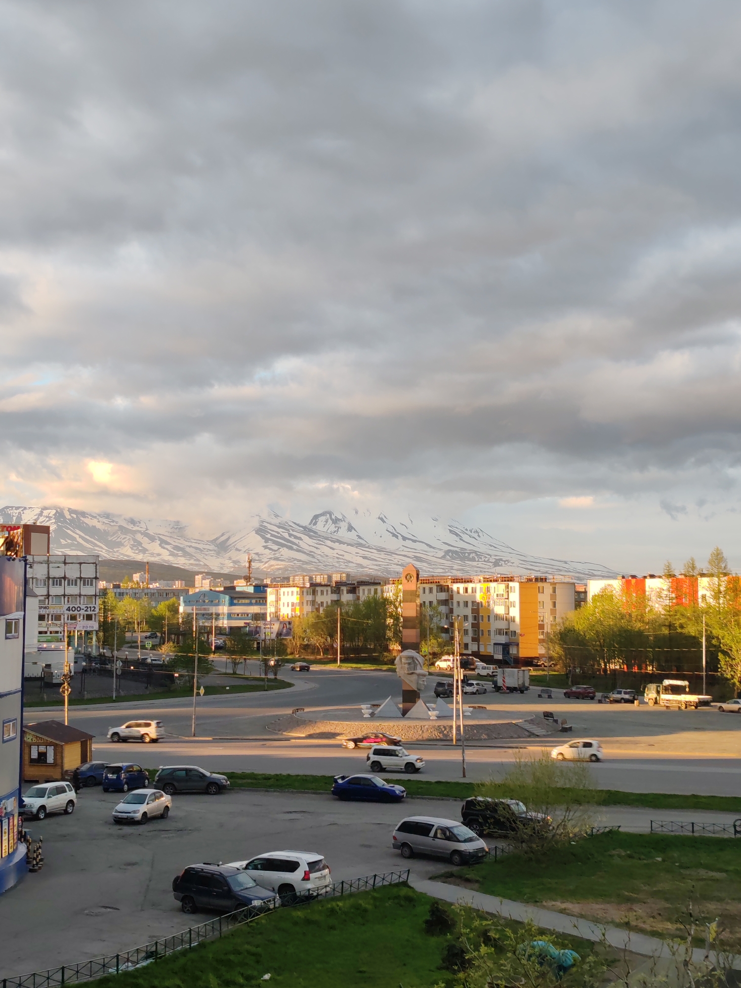 The image depicts a street scene in Petropavlovsk-Kamchatsky, Russia, with snow-capped mountains in the background under a cloudy sky. In the foreground, several cars are parked and driving on a road. Buildings, including apartment blocks and a small kiosk, are visible. Green trees line the street and a grassy area with a fence is also present. The lighting suggests it is either early morning or late afternoon.