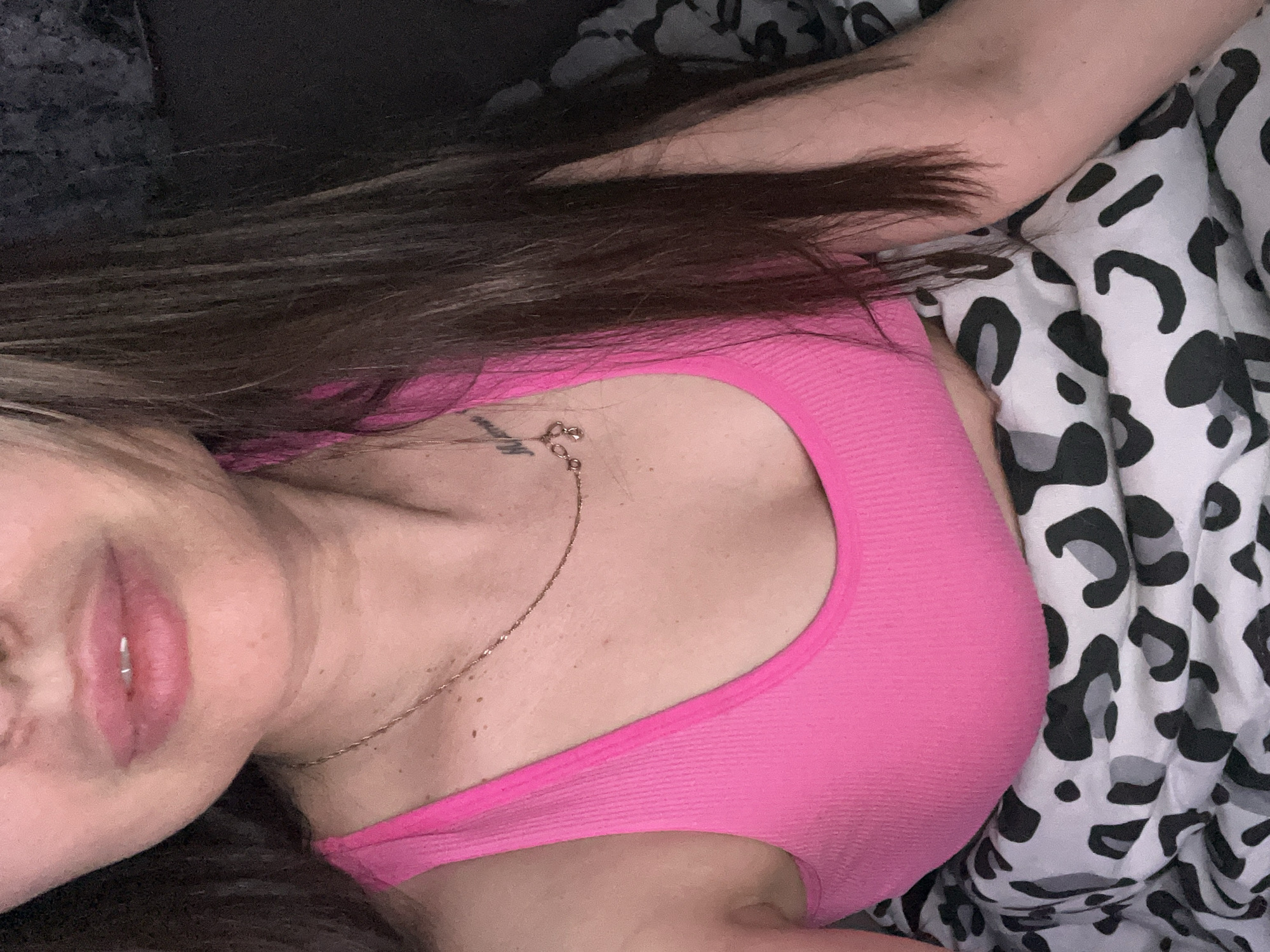 The image shows a woman lying down. She has brown hair and is wearing a bright pink tank top. She has a necklace with a small charm and a lip ring. She is lying on bedding with a black and white animal print. The location is unknown.