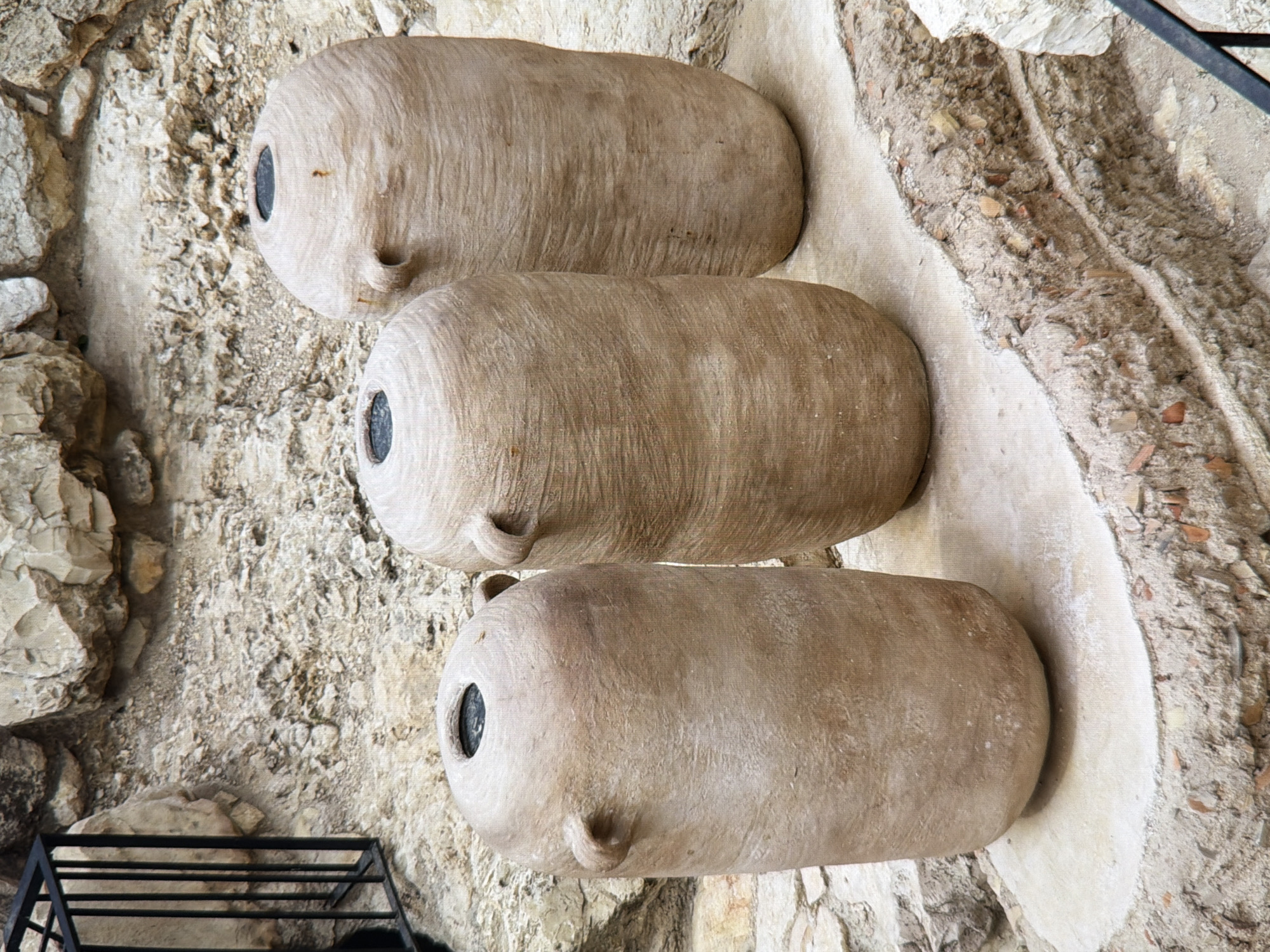 The image shows three large, ancient-looking terracotta amphorae standing upright on a stone surface. The amphorae are beige and have a rough, textured surface. They all have two handles near the neck and a circular opening at the top. They are arranged in a row, with the one on the left slightly in front of the middle one, and the one on the right slightly behind and to the right of the middle one. The background consists of rough, uneven stone walls and earth, suggesting an archaeological dig or an ancient site. There is a black metal railing on the far left of the image. The scene is outdoors, under natural light. There are no people visible in the image. The location is not in Kyiv, Ukraine; the description provided is based solely on the visual information in the image.