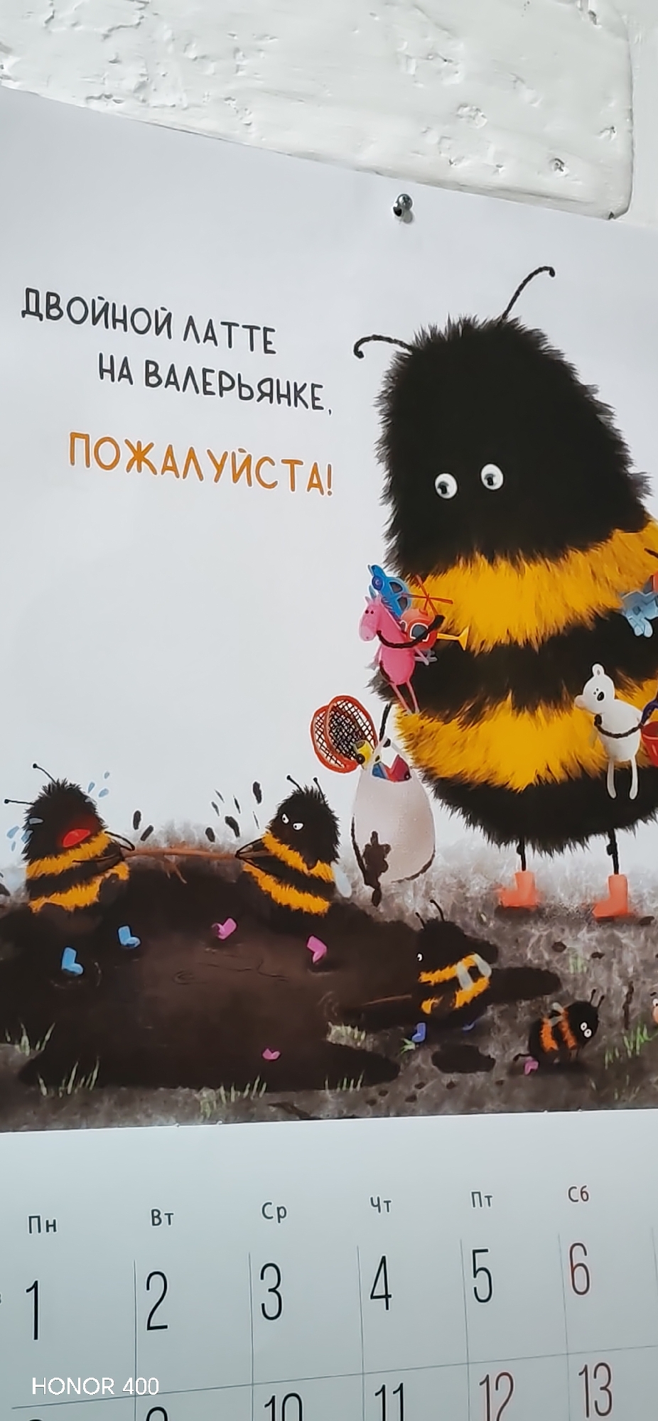 The image displays a close-up of a calendar page, featuring a whimsical illustration, affixed to a textured white wall in Noviy, Russia. The central illustration depicts a large, fluffy, black and yellow striped bumblebee-like creature, likely a parent, looking visibly overwhelmed with wide, stressed eyes. It holds an assortment of colorful toys, including a pink unicorn, a blue helicopter, a tennis racket with a ball, and a white dog-like figure.
Below, several smaller, similarly striped bumblebee-like creatures, presumably children, are shown gleefully playing in a large, dark brown mud puddle, creating splashes. One small bee is crying, another has a mischievous expression, and a third is emerging from the mud, all wearing tiny pink and blue boots. This scene vividly captures the energetic and messy nature of children's play contrasted with parental exhaustion.
Above the illustration, bold Russian text reads: "ДВОЙНОЙ ЛАТТЕ НА ВАЛЕРЬЯНКЕ, ПОЖАЛУЙСТА!" which translates to "DOUBLE LATTE WITH VALERIAN, PLEASE!" This text humorously reinforces the parent's evident need for calm.
The bottom section of the image displays the calendar grid, showing the first few days of a month. Russian abbreviations for the days of the week are visible: Пн (Monday), Вт (Tuesday), Ср (Wednesday), Чт (Thursday), Пт (Friday), and Сб (Saturday). The dates 1, 2, 3, 4, 5, and 6 are clearly marked, with the number 6 highlighted in red, typically indicating a weekend or holiday. Partial dates 10, 11, 12, and 13 are also visible below. A "HONOR 400" watermark is present in the lower left corner, indicating the device used to capture the photograph.