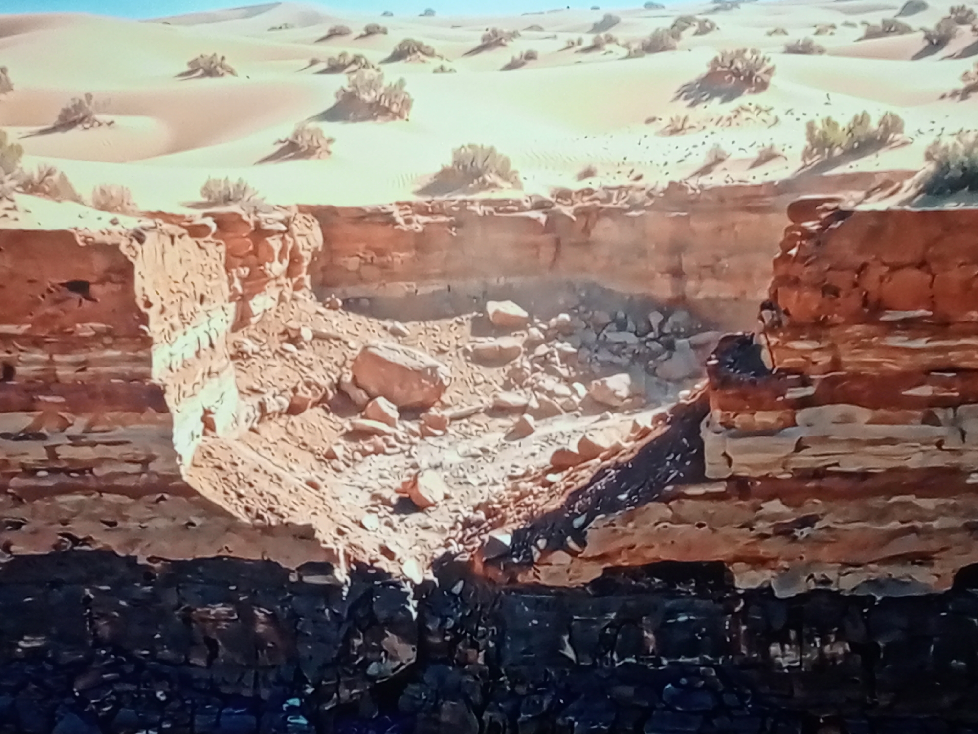 The image depicts a desert landscape with rolling sand dunes dotted with sparse vegetation. In the foreground, a dramatic cliff face has collapsed, revealing layers of sedimentary rock. The eroded cliff has created a large, irregular cavity, with loose rocks and debris scattered at its base. The rock layers exhibit a gradient of colors, from light tan and orange at the top to dark, almost black layers at the bottom. The overall scene suggests a natural erosion event in a dry, arid environment.
